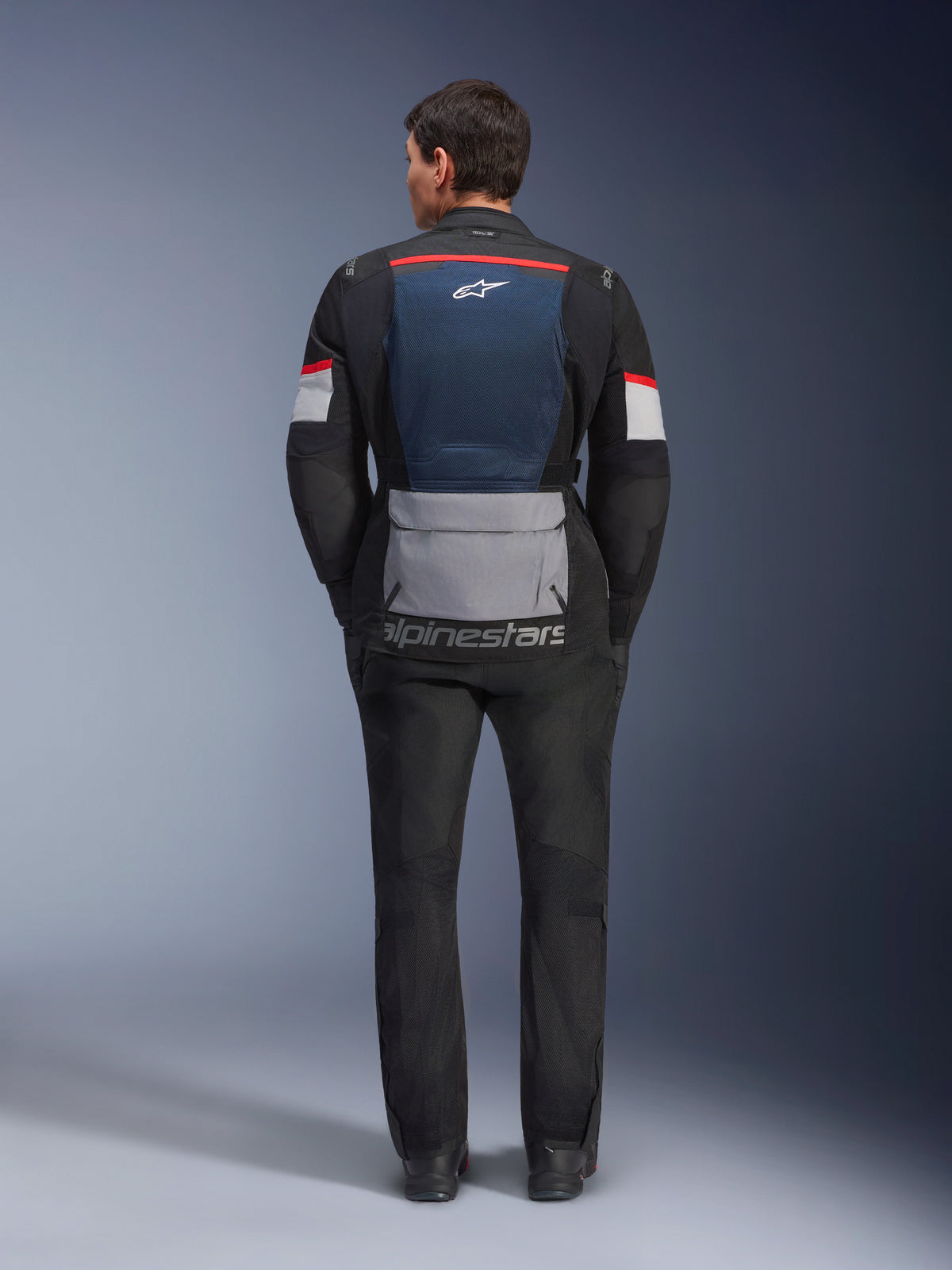 Giacca Alpinestars Andes Air Drystar, Deep Blue Black Ice Gray, vista posteriore su modello, con pannello posteriore in rete blu per la ventilazione, ampia tasca inferiore grigia, dettagli rossi e bianchi sulle maniche, marchio 'alpinestars', abbinata a pantaloni da moto neri per un completo da touring