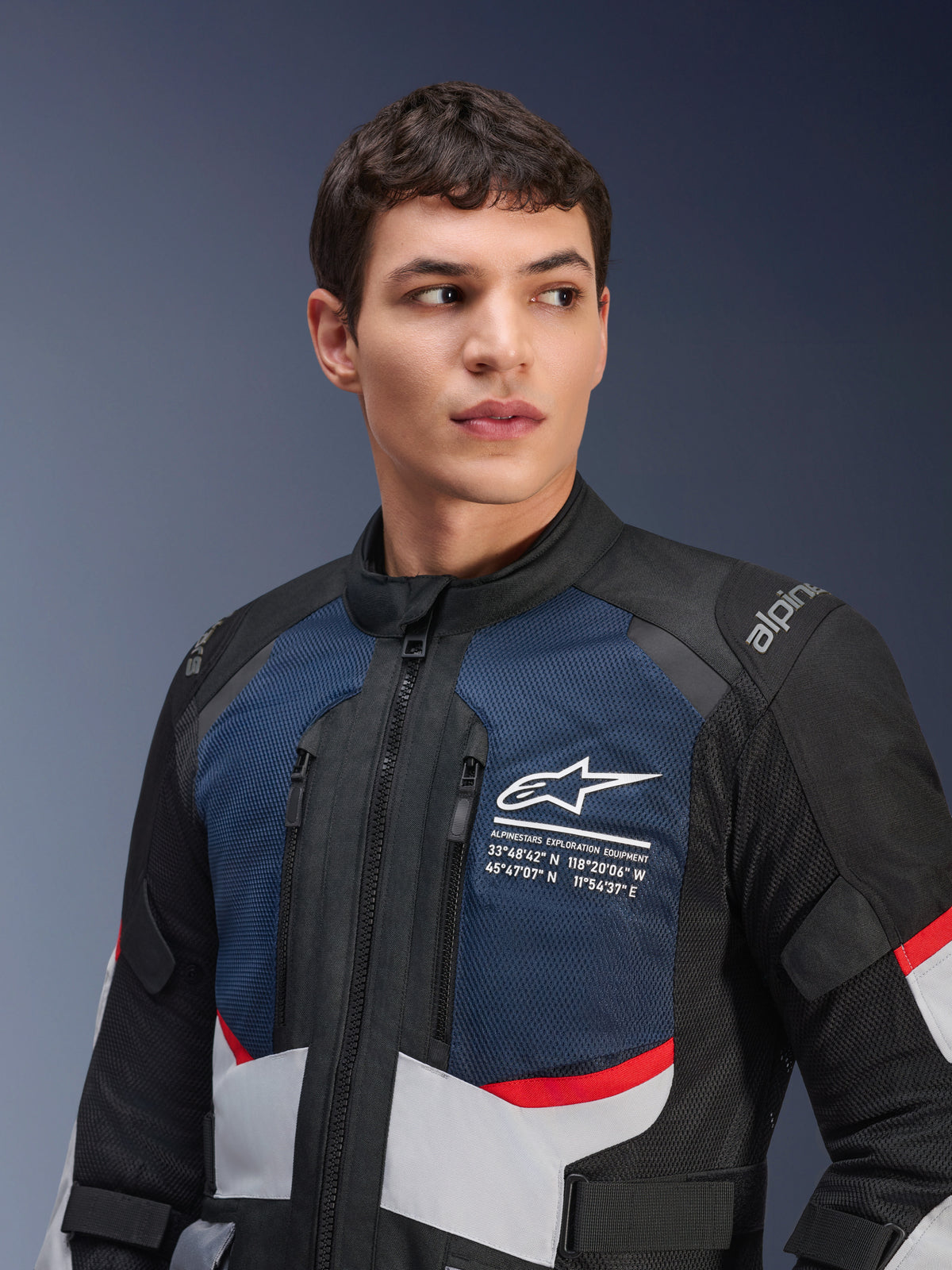 Giacca Alpinestars Andes Air Drystar®, giacca da moto, blu scuro, nero e grigio ghiaccio con accenti rossi, indossata da un modello, con pannelli in mesh ventilata sul petto, logo a stella bianco con coordinate geografiche e design adventure