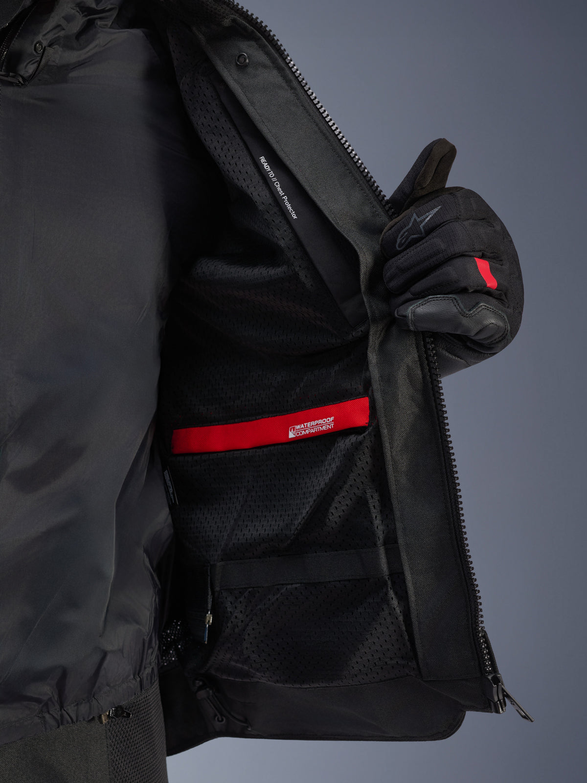 Vista dettagliata dell'interno della giacca Alpinestars Andes Air Drystar®, che mette in evidenza la fodera in rete nera traspirante, lo scomparto impermeabile rosso e la tasca per la protezione del torace, mostrata aperta da un guanto da moto nero.