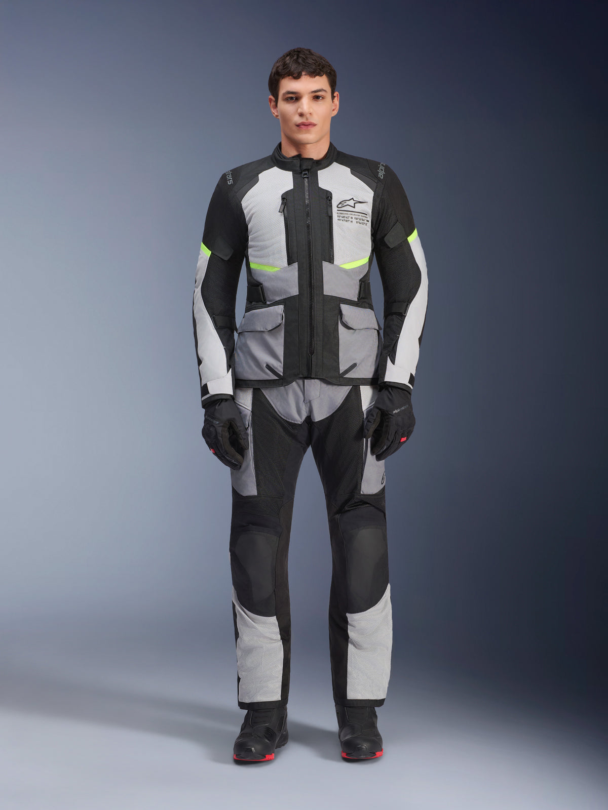 Giacca Alpinestars Andes Air Drystar® in Ice Gray, Dark Gray e Nero, indossata da un modello con pantaloni e guanti da moto abbinati, design adventure touring con grandi tasche anteriori e accenti giallo fluorescente, vista frontale in studio