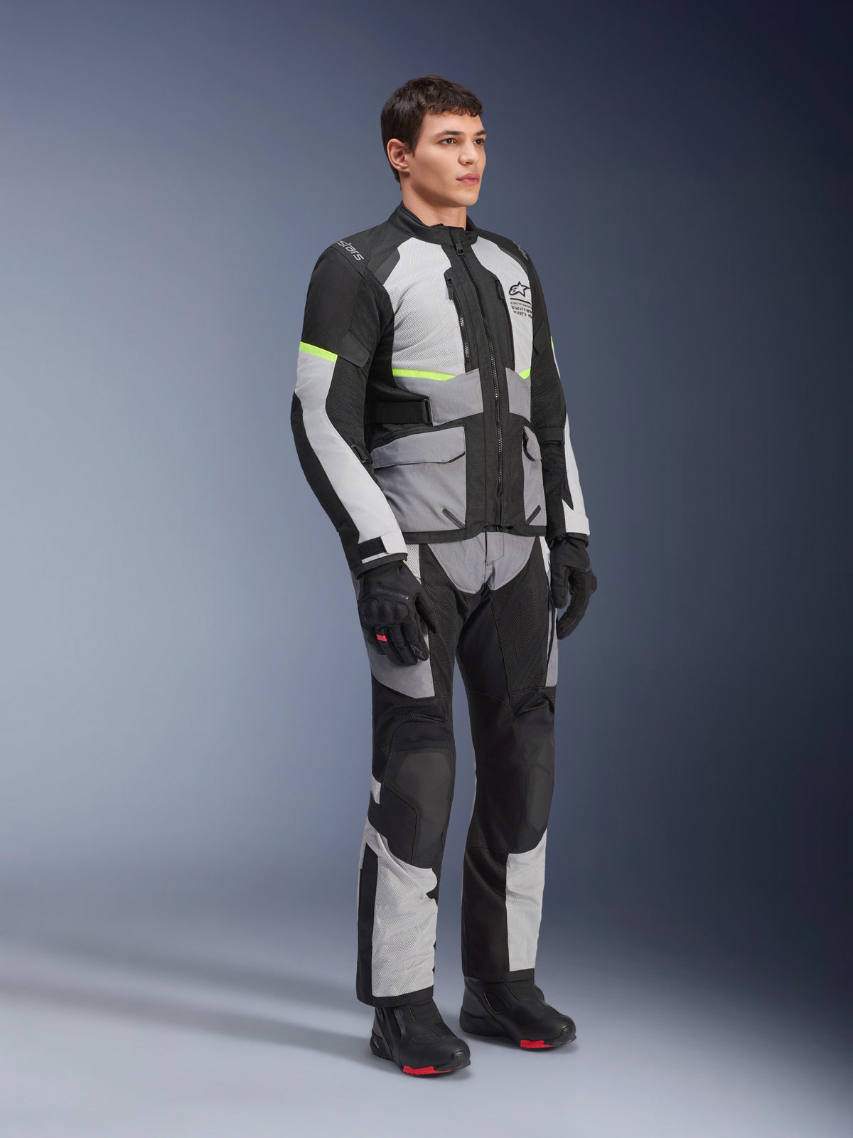 Giacca Alpinestars Andes Air Drystar®, Ice Gray, Dark Gray e nero, indossata da un modello in un outfit completo da adventure touring con pantaloni coordinati e stivali neri, dotata di ventilazione in rete, accenti neon e marchio Alpinestars.