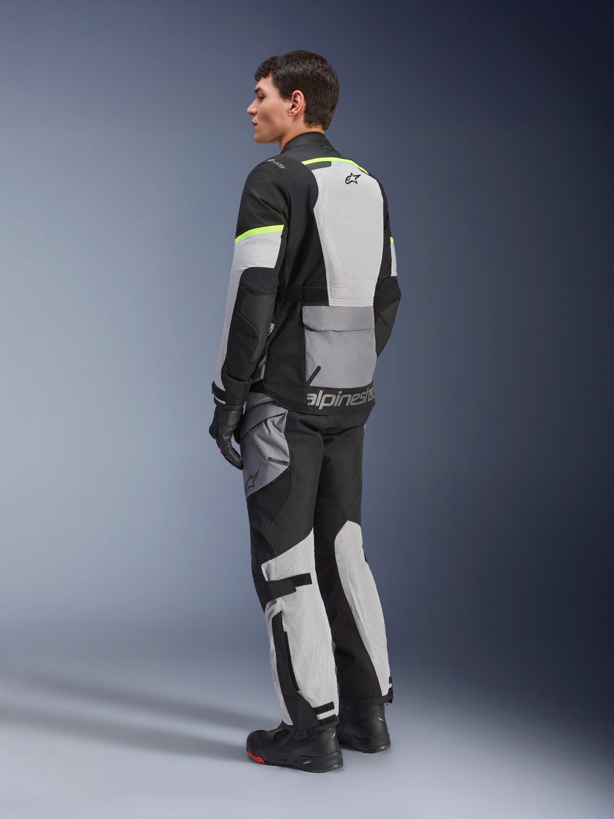 Giacca Alpinestars Andes Air Drystar®, giacca da moto touring, grigio ghiaccio e grigio scuro con pannelli neri e accenti giallo neon, indossata con pantaloni e guanti coordinati, vista posteriore di tre quarti che mostra la ventilazione in rete e il marchio Alpinestars.