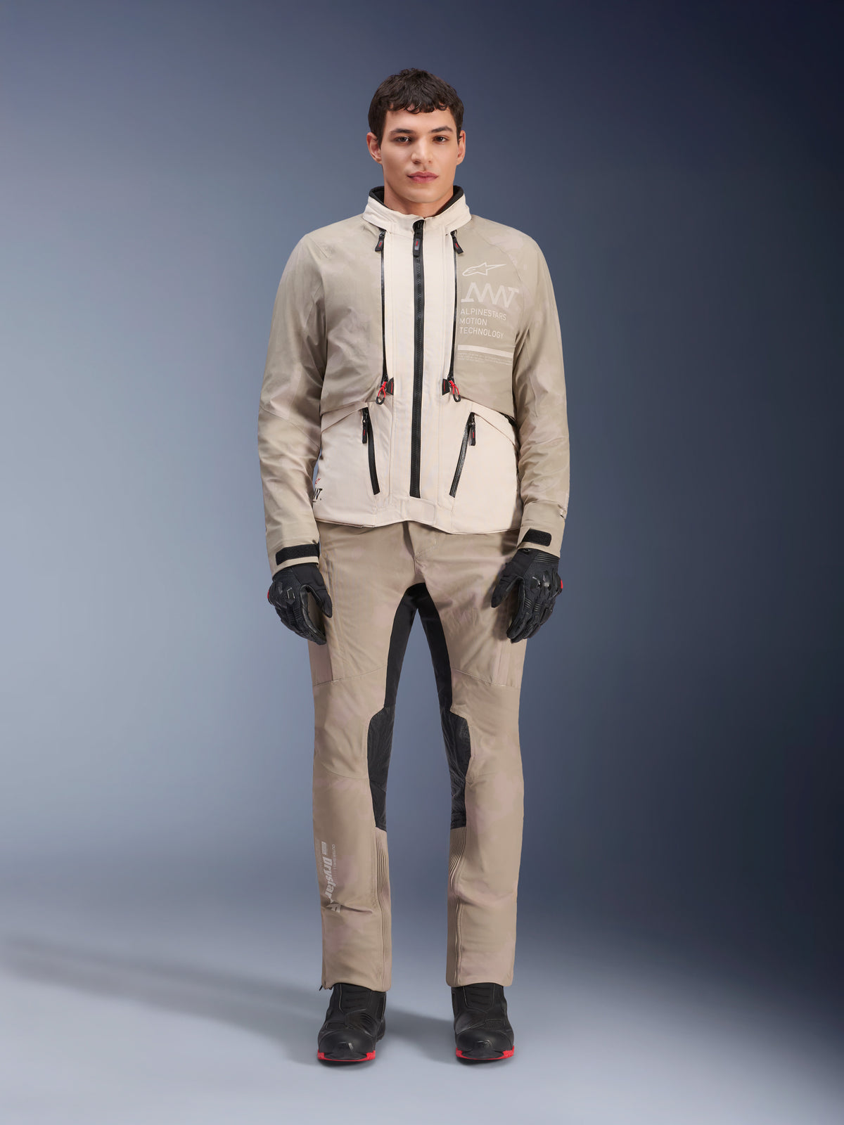 Giacca Alpinestars AMT-10LAB Drystar® XF in White Sand Aluminum Camo, indossata da un modello con pantaloni da moto adventure coordinati, guanti neri e stivali neri, abbigliamento tecnico modulare da moto per touring multi-stagionale.