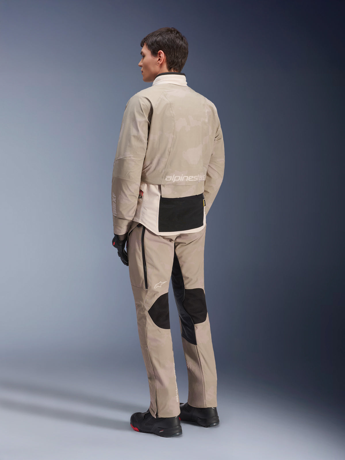 Modello visto di spalle con giacca AMT-10LAB Drystar® XF e pantaloni coordinati, White Sand Aluminum Camo, abbigliamento da moto adventure, con pannelli tecnici neri e logo Alpinestars, indossato con stivali e guanti da moto neri, completo coordinato per il touring in ogni condizione atmosferica