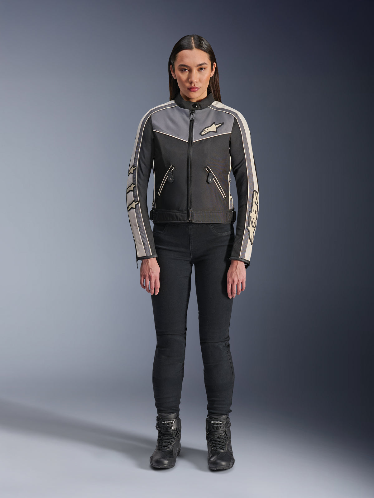 Donna che indossa la giacca Alpinestars Stella T-Dyno WR, Nero Grigio Scuro ed Ecru, vista frontale, giacca da moto in tessuto dal design d'ispirazione vintage, con logo su petto e maniche, indossata con pantaloni e stivali neri