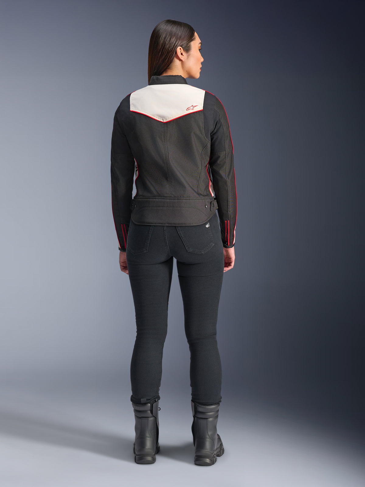 Vista posteriore di una modella che indossa la giacca Alpinestars Stella T-Dyno WR in Nero Ecru Rosso Brillante, caratterizzata da un evidente pannello posteriore color crema e dettagli del logo rossi, abbinata a pantaloni e stivali neri, una giacca da moto in tessuto d'ispirazione vintage.