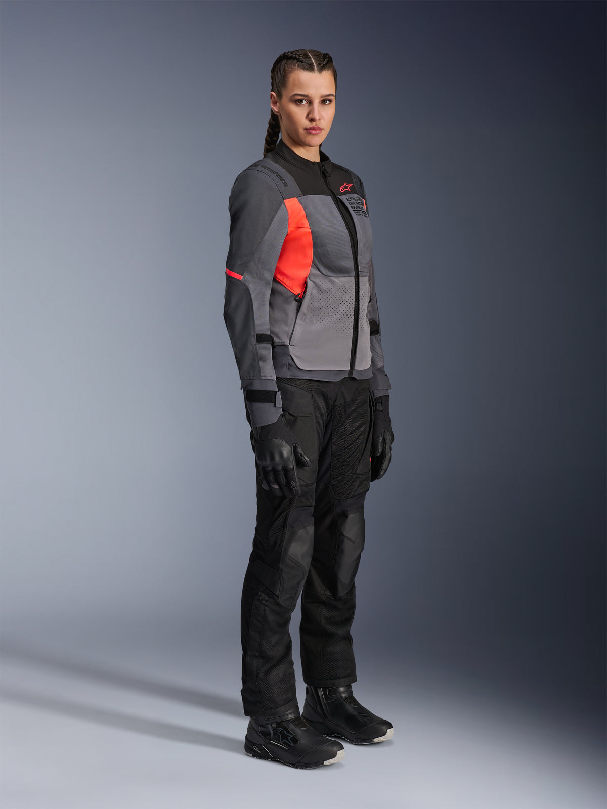 Motociclista donna che indossa la giacca Alpinestars Stella ST-2 Air in Deep Gray, Dark Gray e Fluo Coral, abbinata a pantaloni da moto, guanti e stivali neri, caratterizzata da un design touring moderno con pannelli in mesh e dettagli riflettenti.