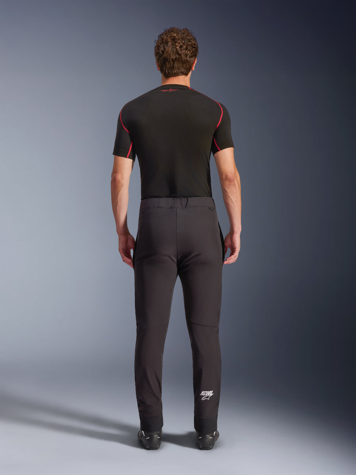 Pantaloni Alpinestars Aeroshell, pantaloni da moto crossover sportivi in softshell, nero, vista posteriore indossati con uno strato base nero a maniche corte, caratterizzati da vestibilità slim, ginocchia pre-curvate e logo Astars bianco sulla parte inferiore della gamba sinistra.