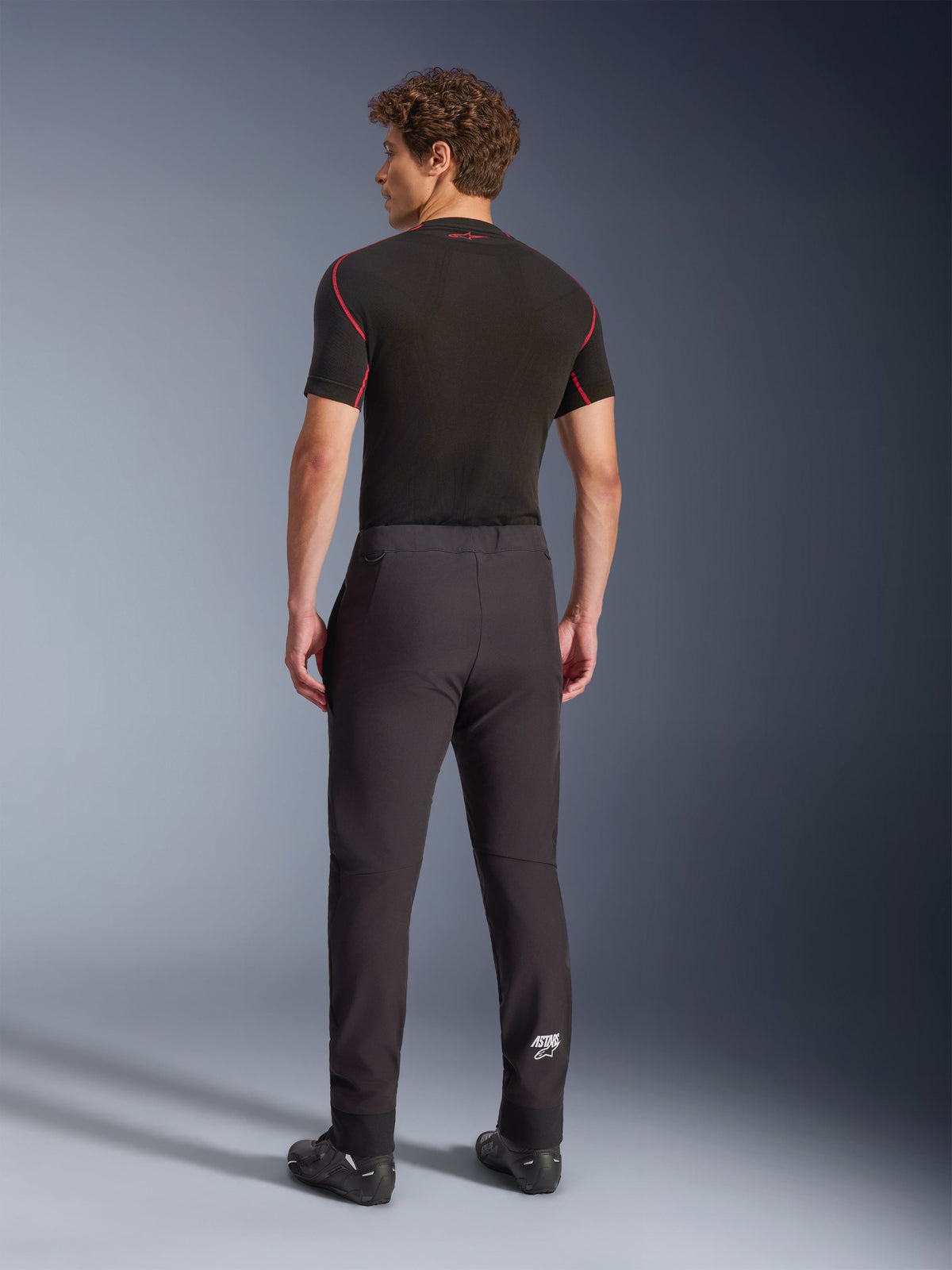 Pantaloni Alpinestars Aeroshell, nero, pantaloni da moto in softshell, vista posteriore su un modello, caratterizzati da un taglio moderno affusolato, logo Astars bianco sulla parte inferiore della gamba e orli elasticizzati, indossati con un base layer nero a maniche corte e scarpe da moto.