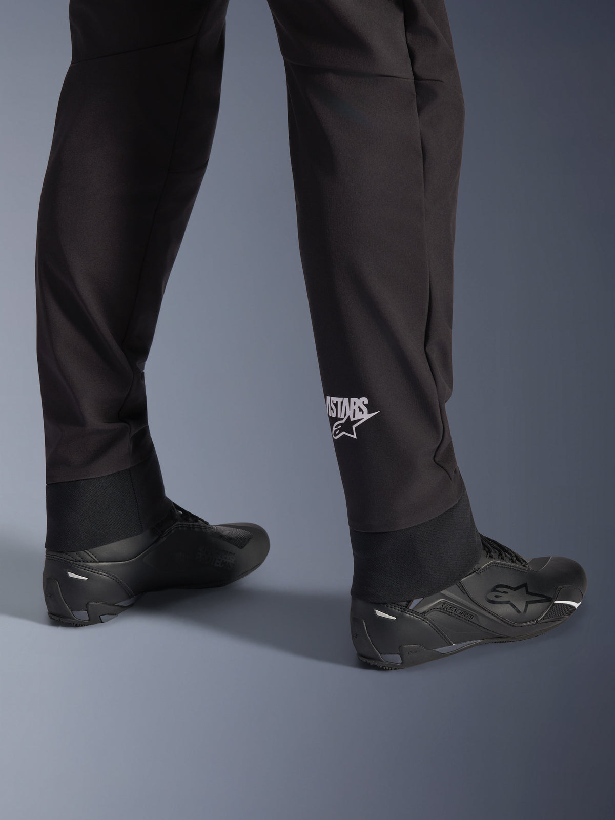 Alpinestars Aeroshell Pants, pantaloni da moto in softshell neri, dettaglio della gamba e dell'orlo elasticizzato, logo Alpinestars bianco sul polpaccio, abbinati a scarpe da moto nere, design crossover sportivo moderno