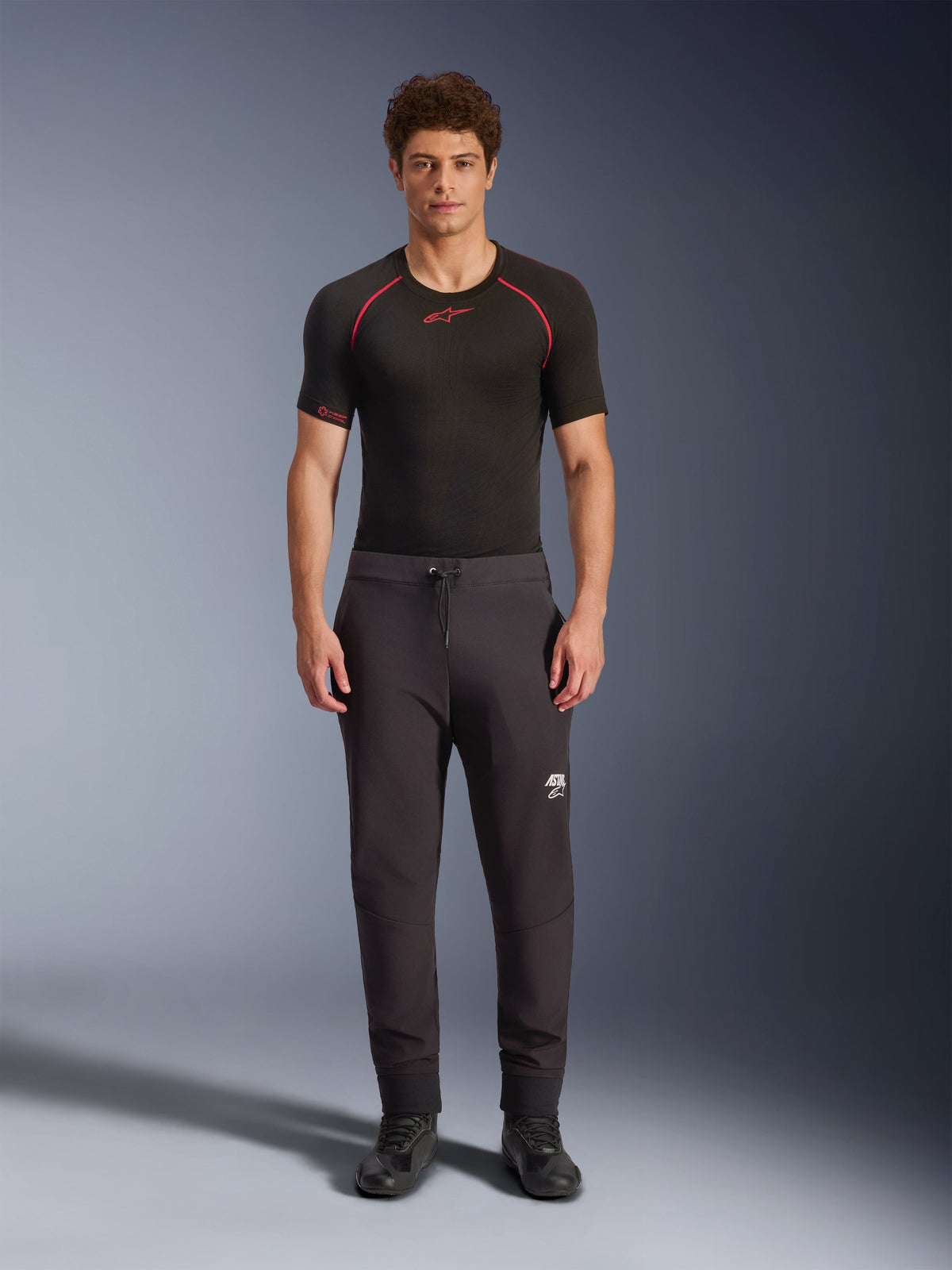 Alpinestars Aeroshell Pants, pantaloni da moto softshell neri, indossati con un base layer nero a maniche corte e scarpe nere, caratterizzati da vita elastica con coulisse, logo Astars bianco sulla coscia e bordi elastici, silhouette atletica moderna per la guida su strada