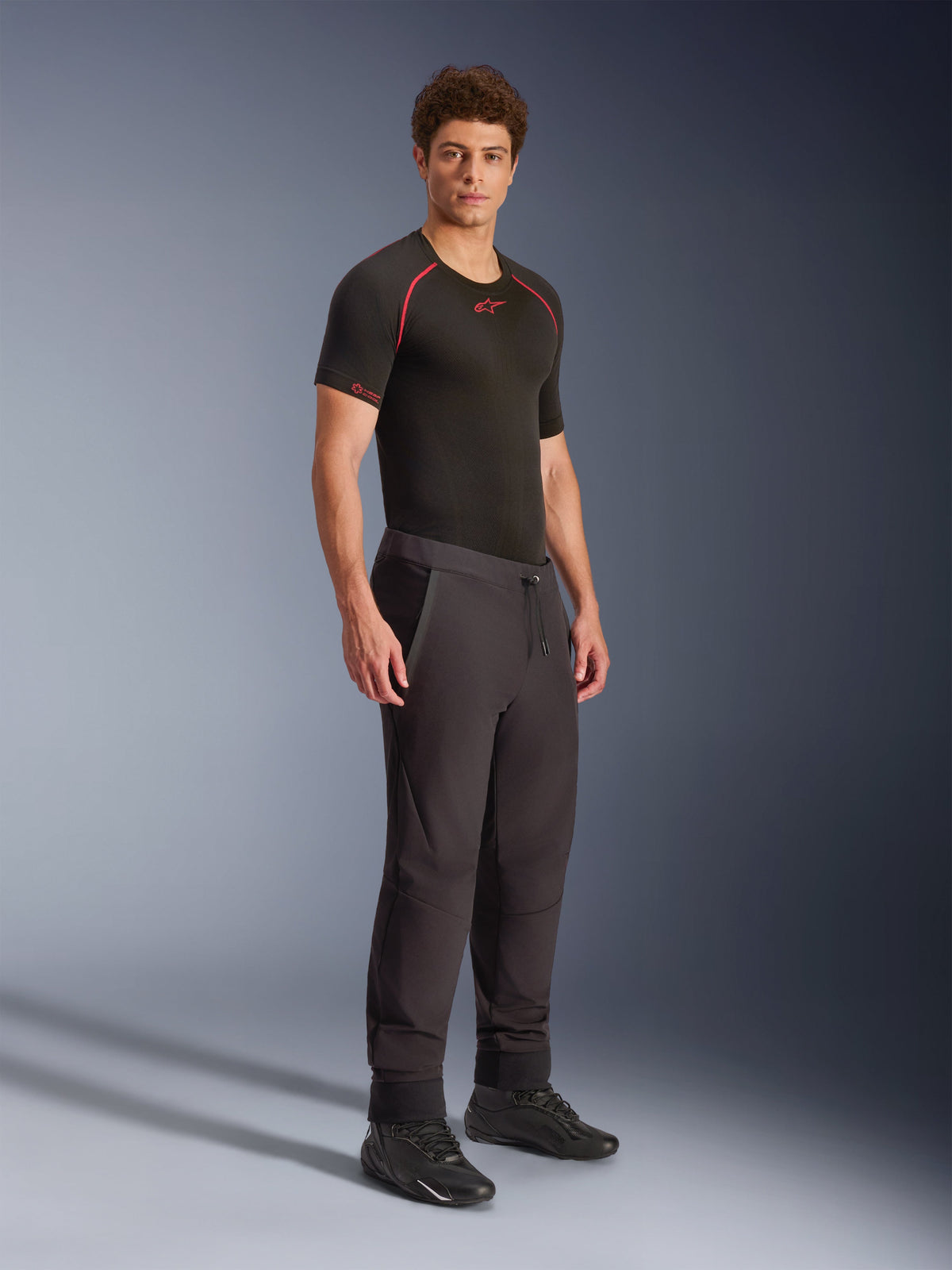 Alpinestars Pantaloni Aeroshell, nero, pantaloni da moto softshell sportivi crossover, indossati da un modello con base layer tecnico nero e scarpe nere, caratterizzati da vita con cordoncino e orli elastici