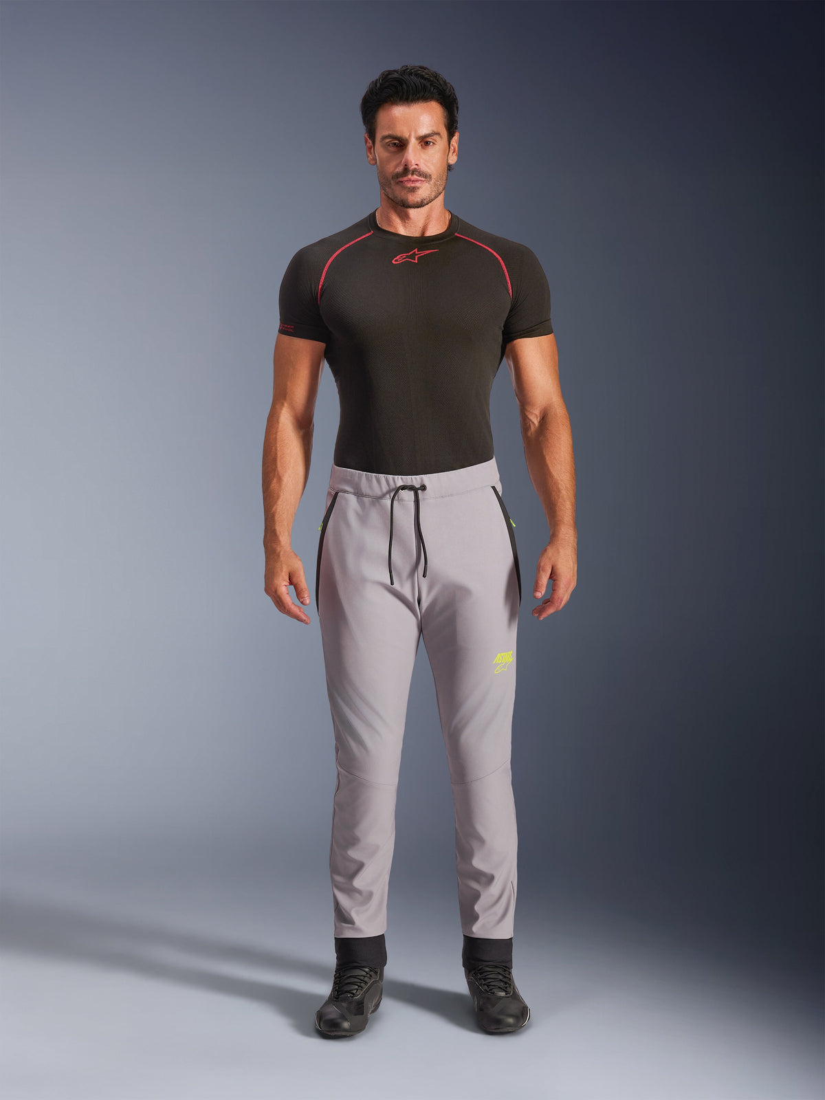 Modello maschile che indossa pantaloni Alpinestars Aeroshell in Grigio Medio e Giallo Fluo, pantaloni softshell sportivi crossover con vita elastica a cordoncino e tasche con zip, abbinati a una maglia tecnica nera a maniche corte, design atletico moderno per la guida su strada e casual