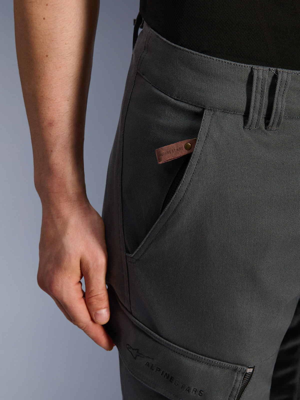 Alpinestars Borrego Drystar® Pants, pantaloni da moto adventure, grigio antracite, dettaglio ravvicinato della tasca e del fianco, texture in tela cerata con etichetta in pelle marrone con logo e marchio Alpinestars ricamato sulla tasca cargo della coscia.