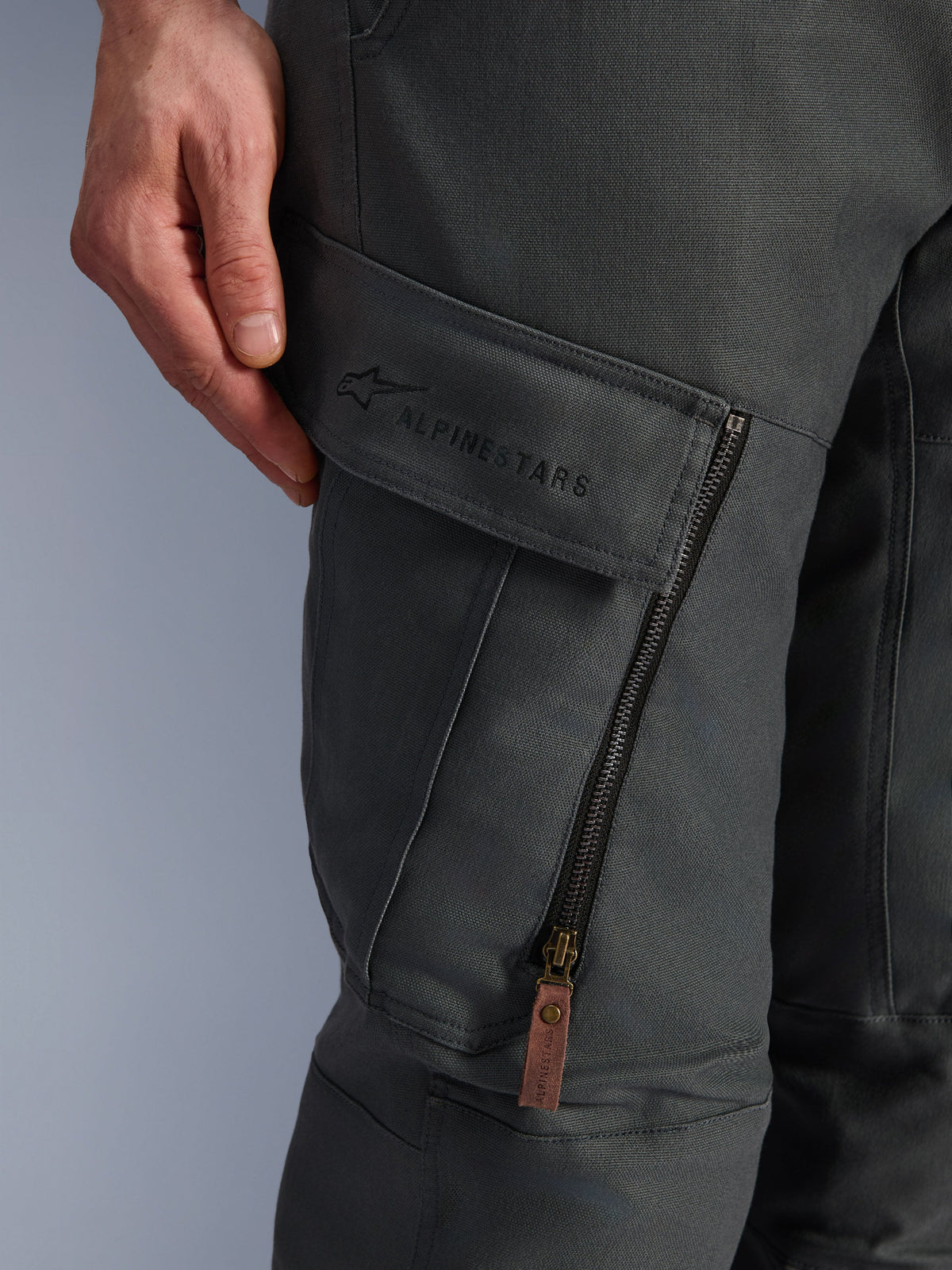 Pantaloni Alpinestars Borrego Drystar®, Grigio Antracite, primo piano della tasca cargo sulla coscia con logo ricamato, materiale in tela cerata testurizzata, cerniera di ventilazione verticale con tirante in pelle marrone