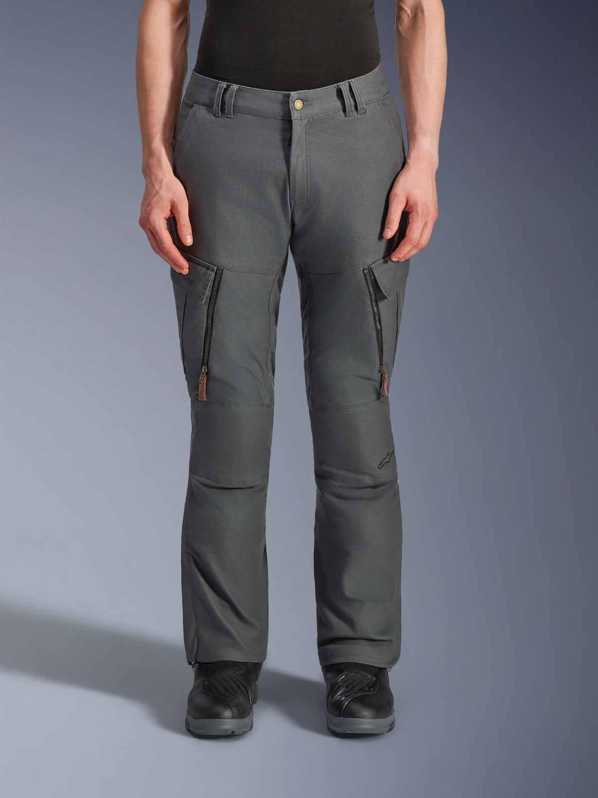 Alpinestars Borrego Drystar® Pants, pantaloni da moto adventure, grigio antracite, con tasche verticali con zip sulle cosce e tiranti in pelle, chiusura della vita con bottone e passanti per cintura, indossati da un modello con stivali da moto neri