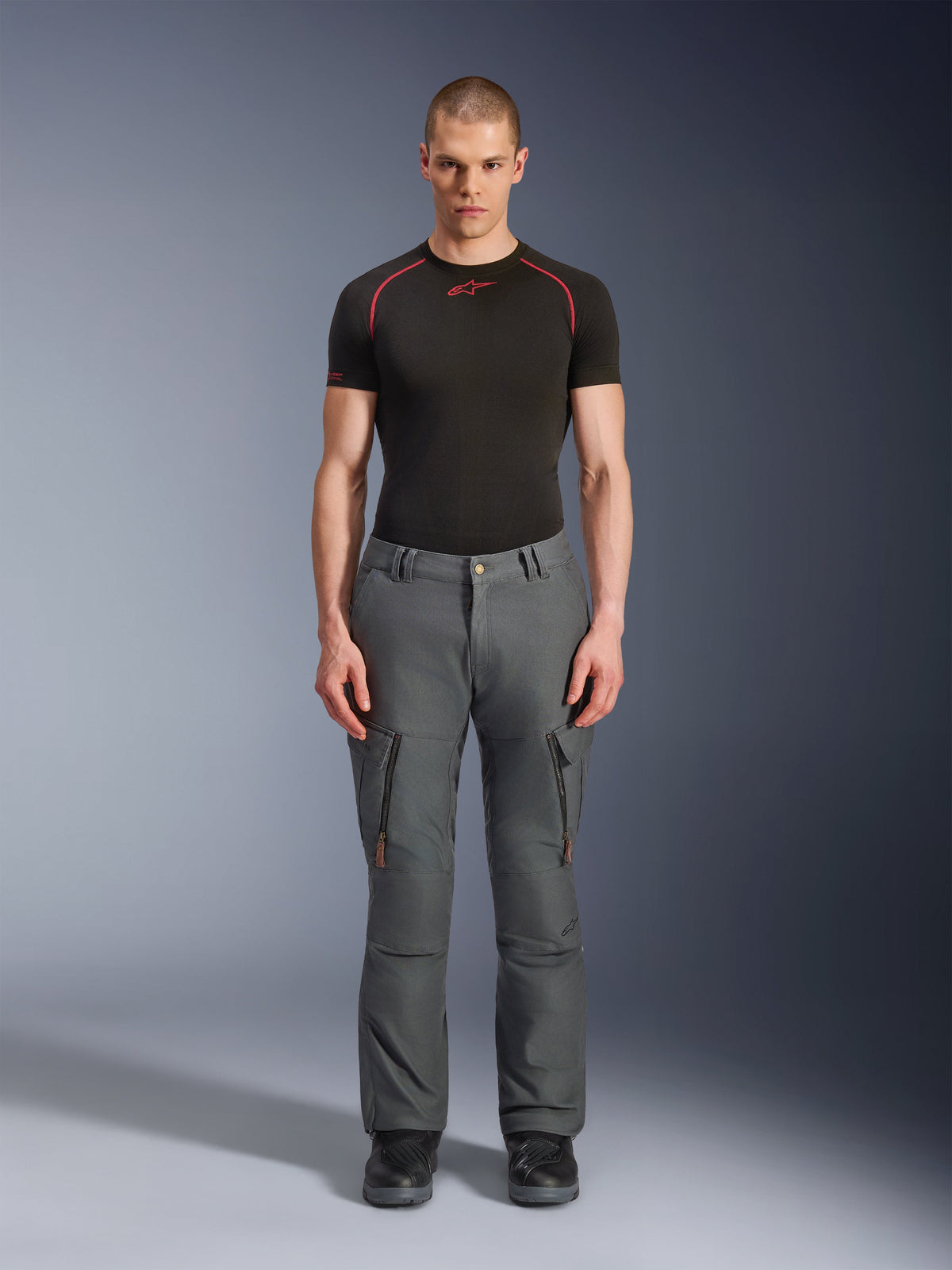 Pantaloni Alpinestars Borrego Drystar®, pantaloni da moto adventure grigi, indossati da un modello con t-shirt tecnica nera e rossa e stivali neri, con tasche cargo dotate di cerniere verticali, aspetto in tela cerata robusta, design resistente per adventure touring e guida su strada