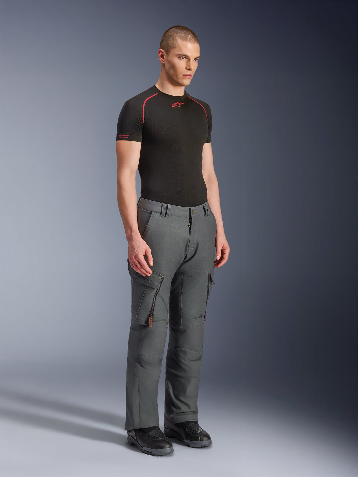 Modello che indossa i pantaloni Alpinestars Borrego Drystar®, pantaloni da moto adventure grigio antracite, design ispirato all'abbigliamento da lavoro con tasche cargo e prese d'aria con zip, abbinati a un base layer nero a maniche corte e stivali neri