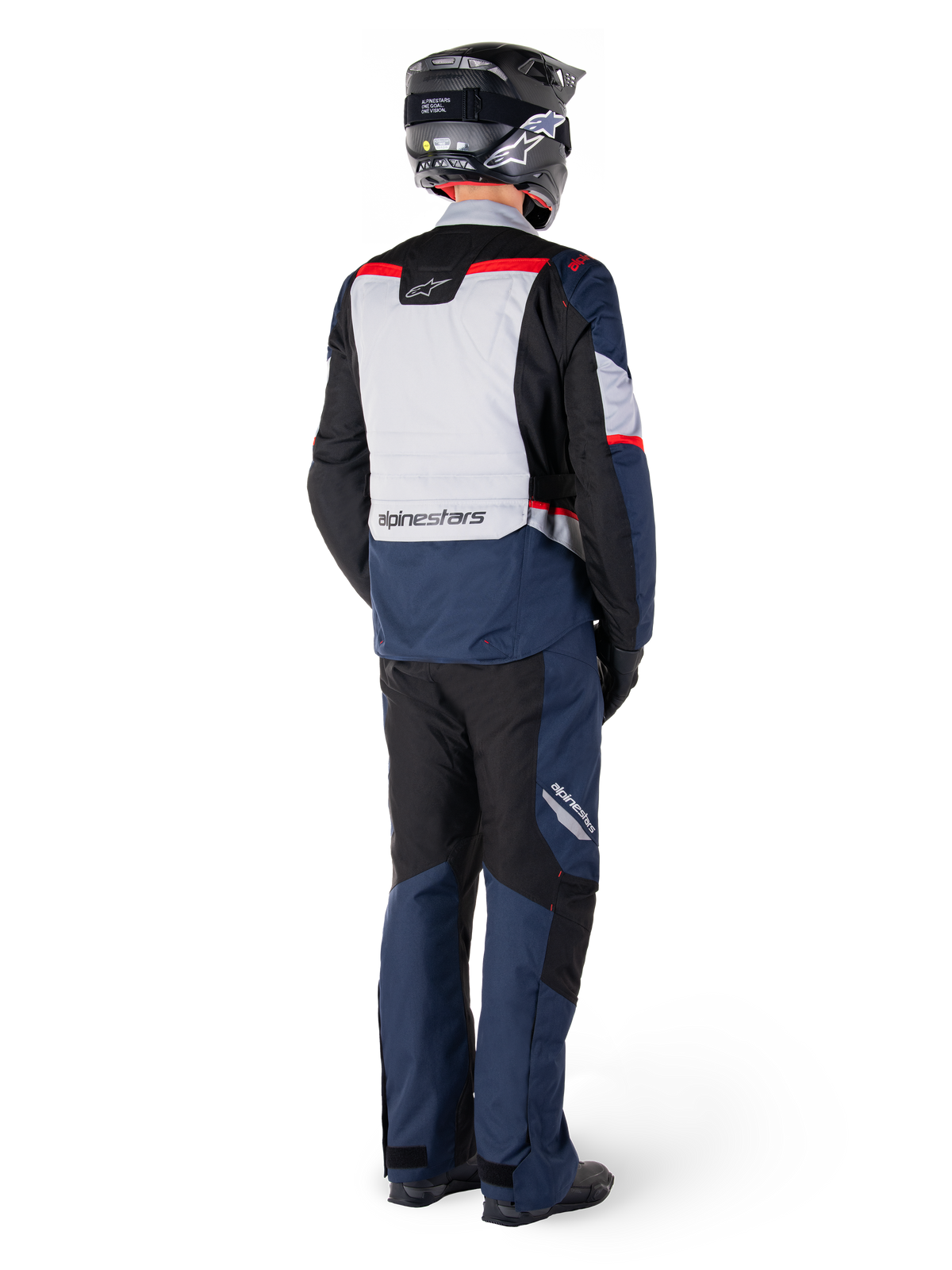 Vista posteriore di un motociclista con pantaloni impermeabili Alpinestars ST-1 e giacca coordinata in blu scuro, nero e rosso brillante, abbigliamento adventure touring con pannelli in tessuto e dettagli riflettenti, casco e stivali neri.