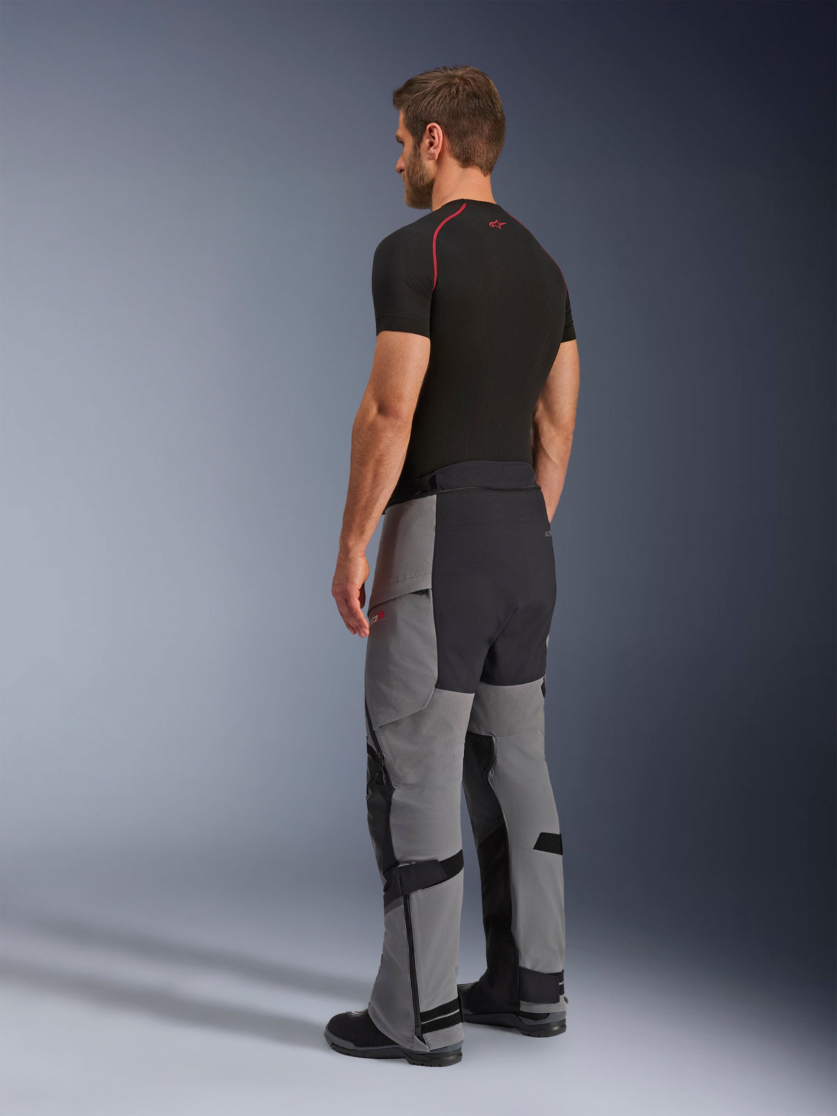 Alpinestars Andes Pro Drystar® XF Pants, pantaloni da moto adventure, grigio scuro e nero, vista posteriore a tre quarti su modello, con costruzione in tessuto tecnico e pannelli rinforzati neri sulla seduta e sull'interno gamba, indossati con una t-shirt base layer nera.