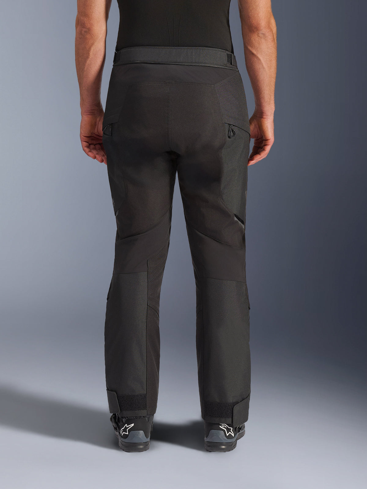 Pantaloni Alpinestars Cusco Drystar®, pantaloni da moto, nero, vista posteriore indossati da un pilota, con pannelli in tessuto testurizzato, prese d'aria con cerniera sulle cosce e chiusure regolabili alle caviglie, abbinati a stivali Alpinestars neri.