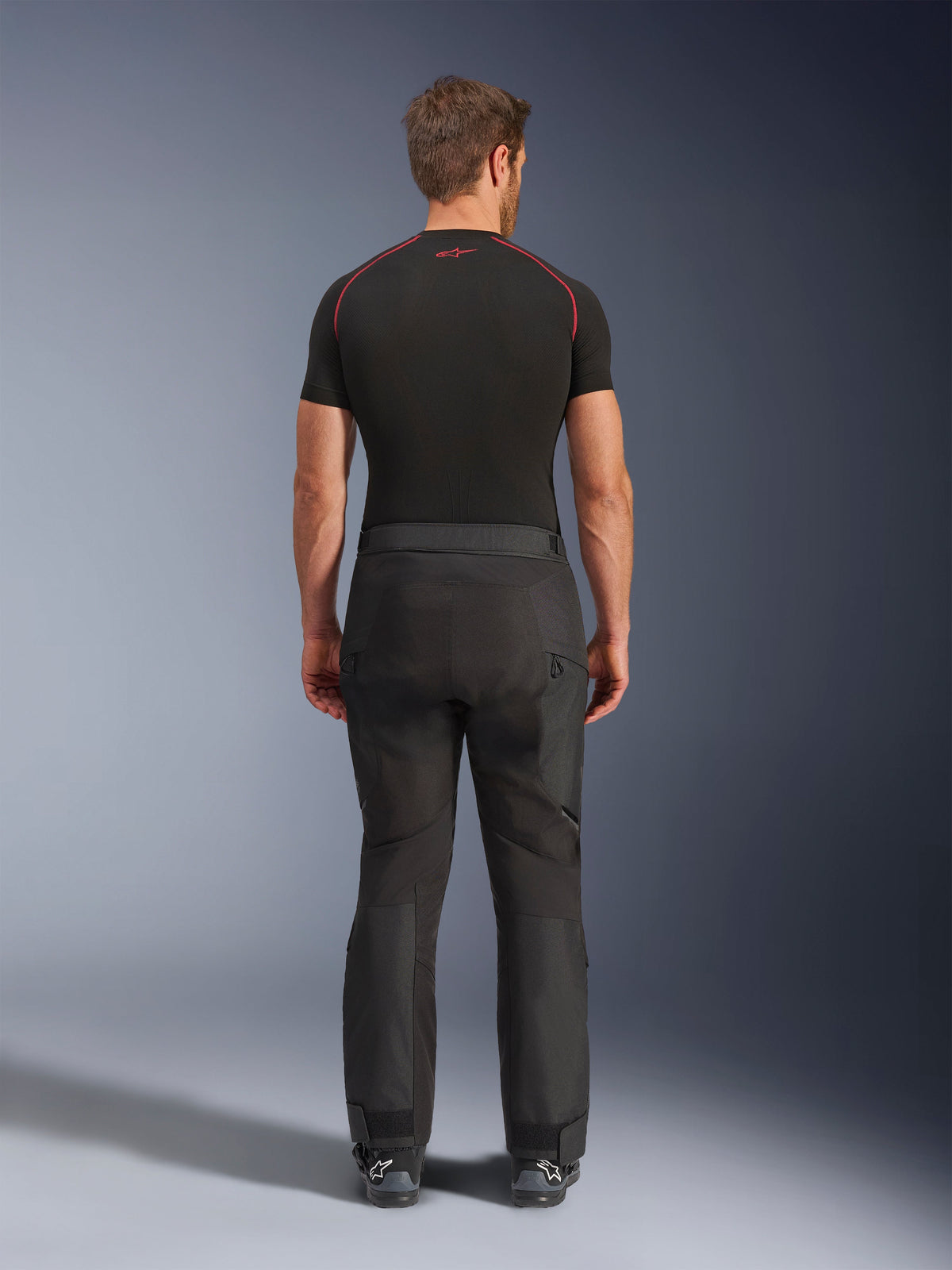 Pilota che indossa i pantaloni Alpinestars Cusco Drystar®, pantaloni da moto adventure neri, vista posteriore, abbinati a una maglia a compressione nera a maniche corte e stivali da moto neri, caratterizzati da un design in tessuto resistente con pannelli articolati per il touring