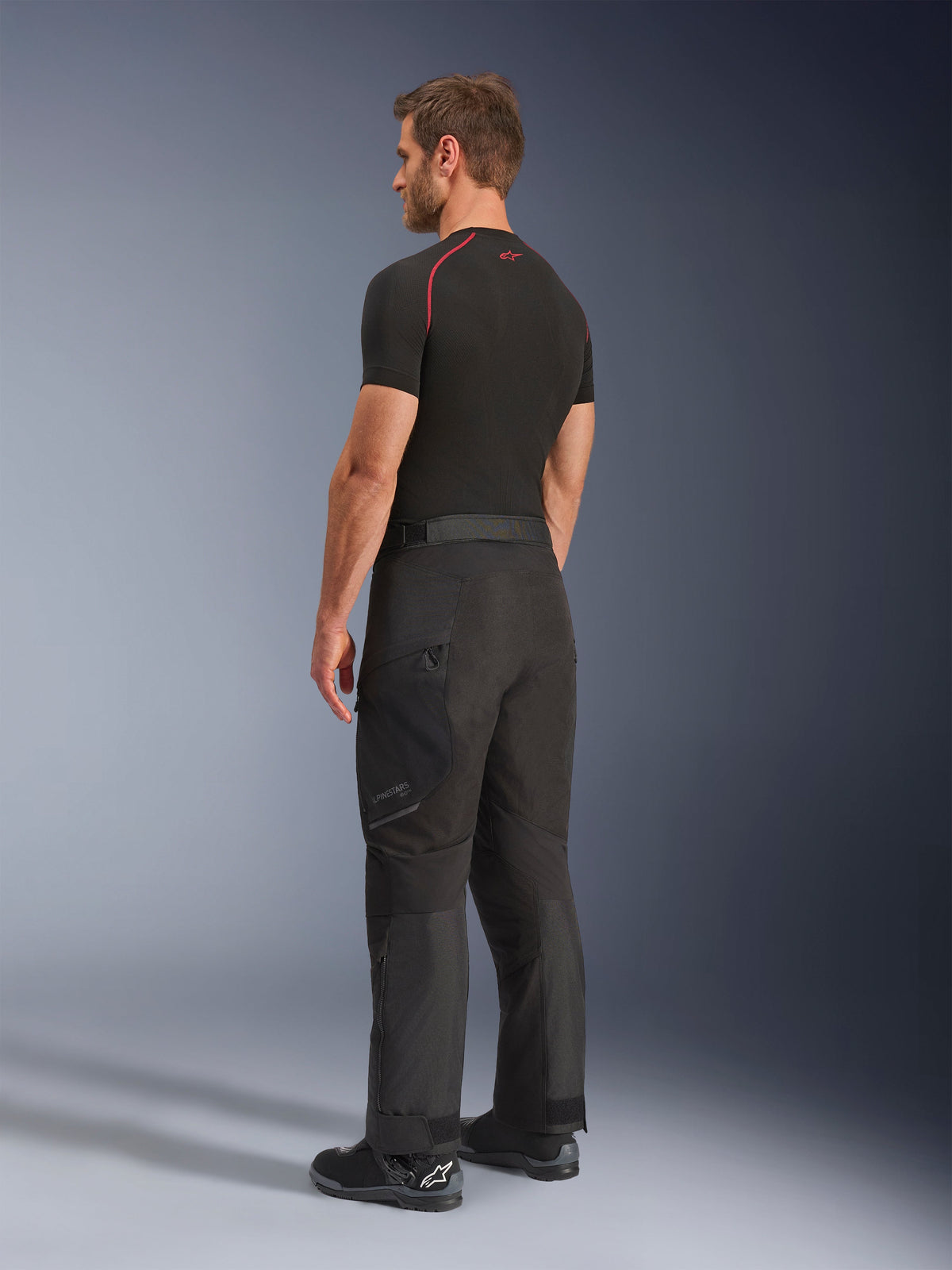 Pantaloni Alpinestars Cusco Drystar®, pantaloni da moto adventure, nero, indossati da un modello in una vista laterale e posteriore, abbinati a un base layer nero e stivali da moto neri, costruzione in tessuto tecnico con tasche con cerniera e ginocchia articolate