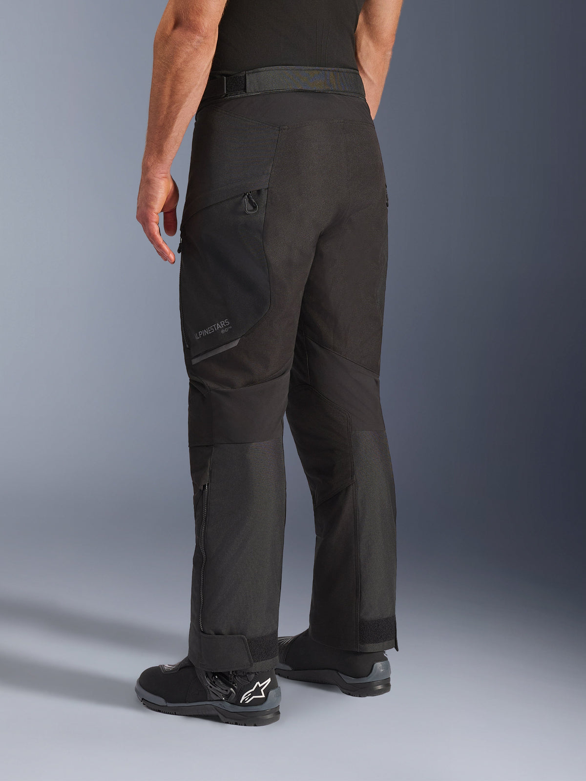 Pantaloni Alpinestars Cusco Drystar®, pantaloni da moto, nero, vista posteriore a tre quarti che mostra la tasca sulla coscia con cerniera e i pannelli articolati, indossati con stivali da moto neri, design resistente per adventure touring con branding discreto