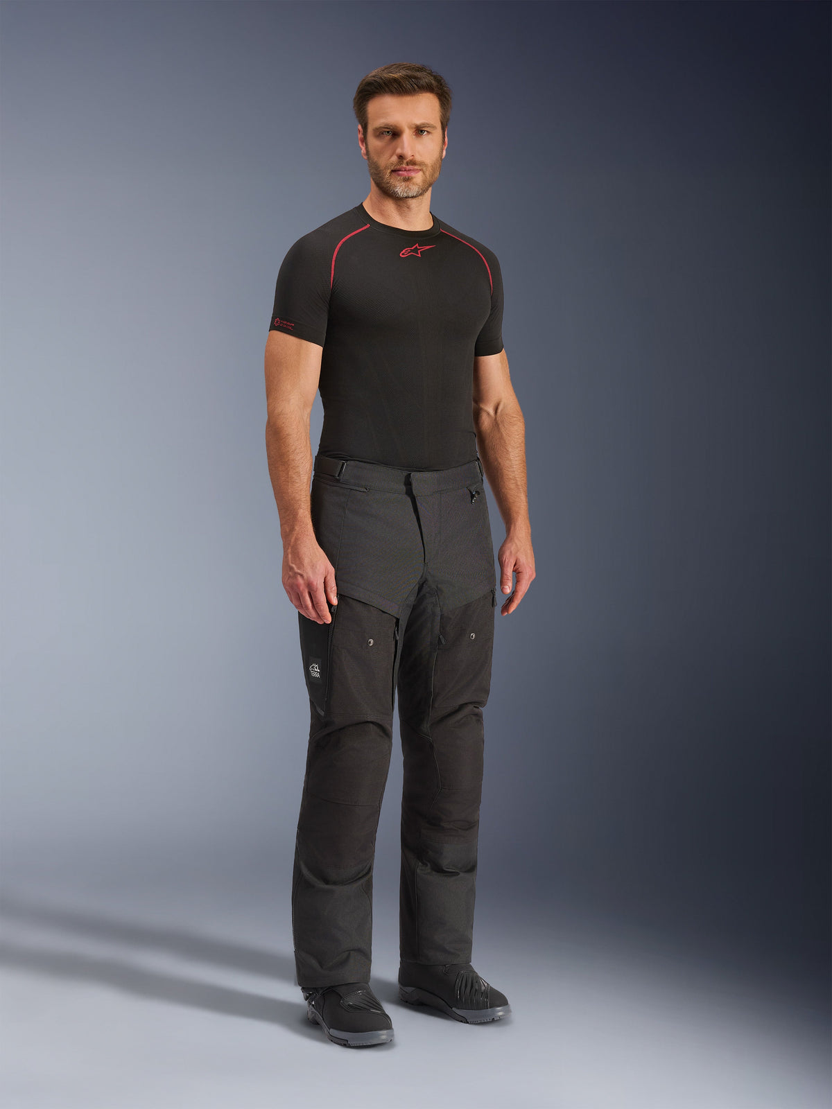 Pantaloni Alpinestars Cusco Drystar®, pantaloni da moto in tessuto nero, indossati da un modello con base layer nero e stivali, caratterizzati da tessuto ripstop, ampie tasche cargo con marchio 'Terra' e design ergonomico per la guida adventure.