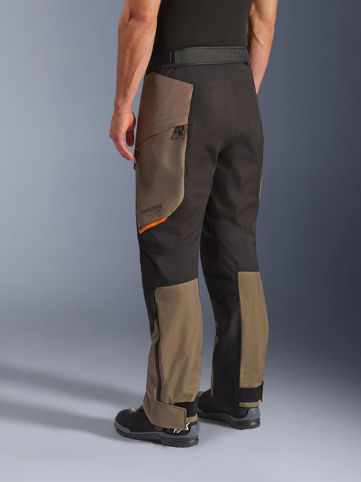 Alpinestars Cusco Drystar® Pants, pantaloni da moto adventure, colore Walnut Black con un piccolo accento arancione, vista posteriore a tre quarti che mostra la costruzione in tessuto e i pannelli color noce, indossati con stivali da moto neri.