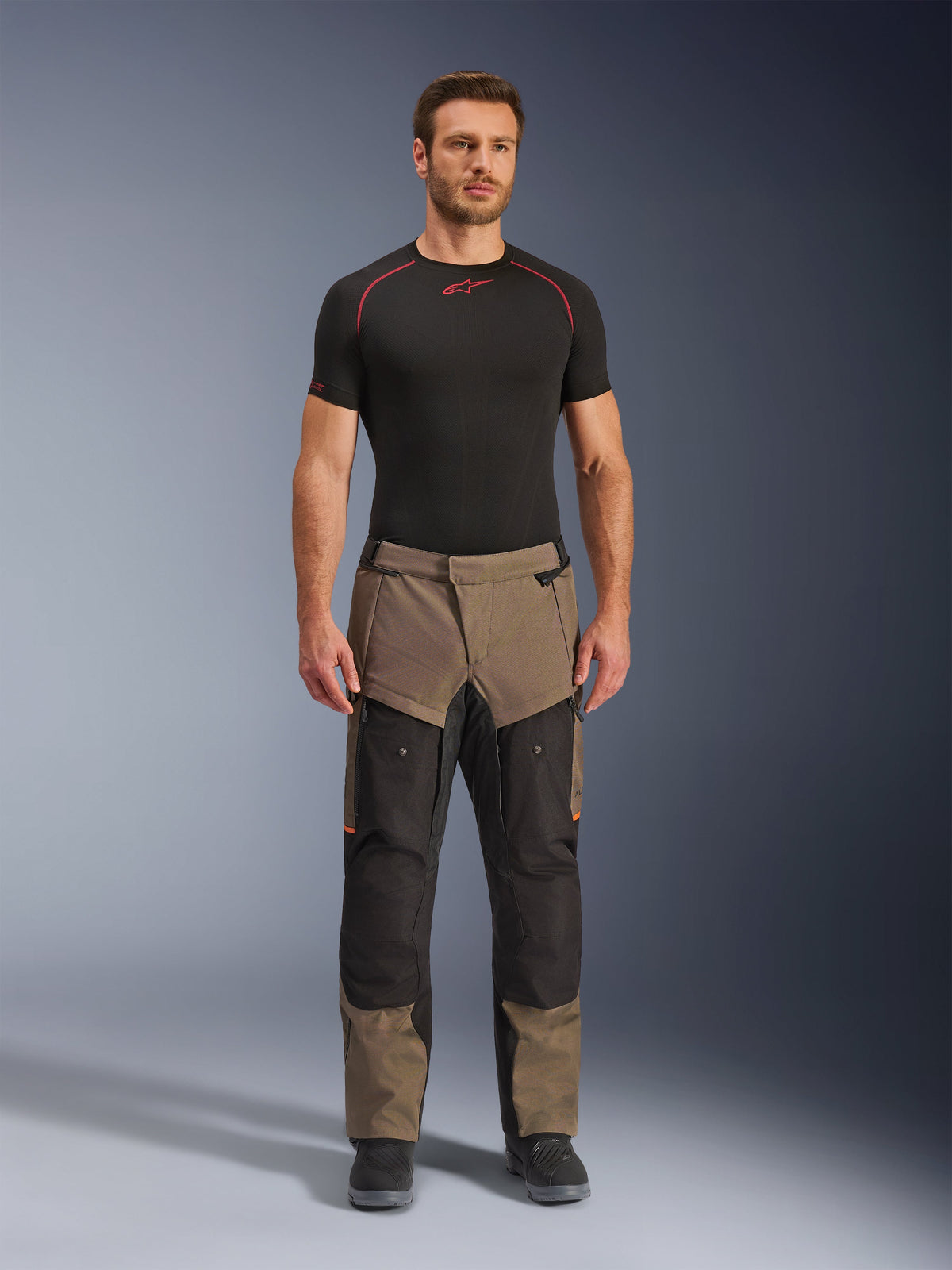 Pantaloni Alpinestars Cusco Drystar®, Walnut Black, pantaloni da moto adventure touring, indossati da un modello con base layer nero e stivali neri, con pannelli neri e color noce, dettagli arancioni e cerniere di ventilazione sulle cosce.