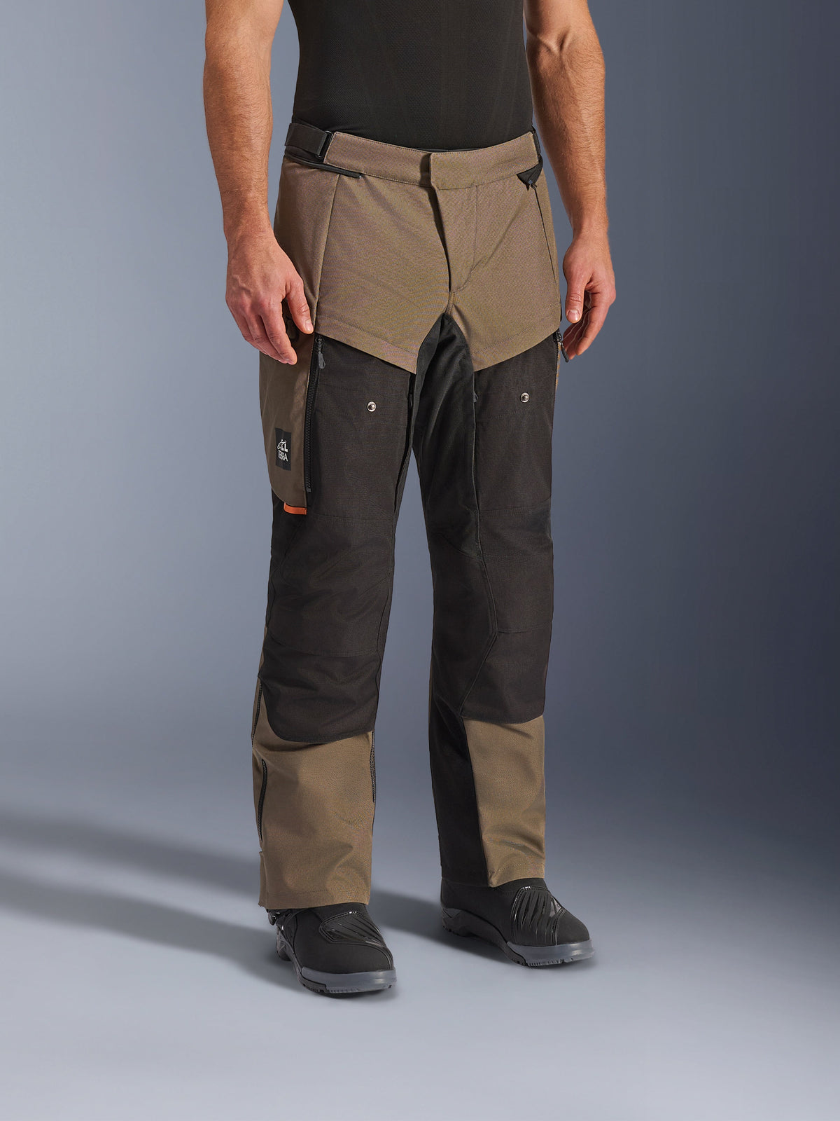 Alpinestars Cusco Drystar® Pants, pantaloni da moto adventure, Walnut Black, indossati da un modello con stivali neri, caratterizzati da un design a pannelli multipli marrone e nero con tasche con cerniera e ginocchia rinforzate per il touring