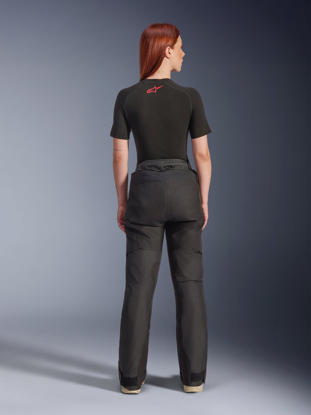 Vista posteriore di una modella che indossa i pantaloni Alpinestars Stella Andes V4 Drystar® neri, abbinati a un base layer nero a maniche corte con logo Alpinestars rosso, abbigliamento da moto adventure-touring con struttura in tessuto resistente.