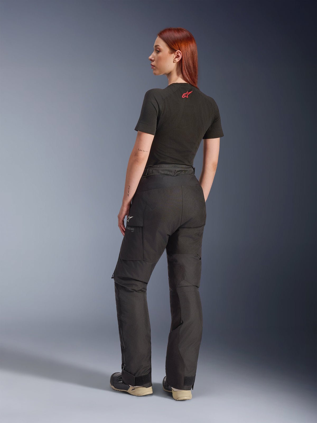 Pantaloni Alpinestars Stella Andes V4 Drystar®, neri, pantaloni da moto adventure, vista laterale posteriore su una modella, abbinati a t-shirt nera e stivali, tasche cargo laterali con logo bianco e struttura in tessuto resistente