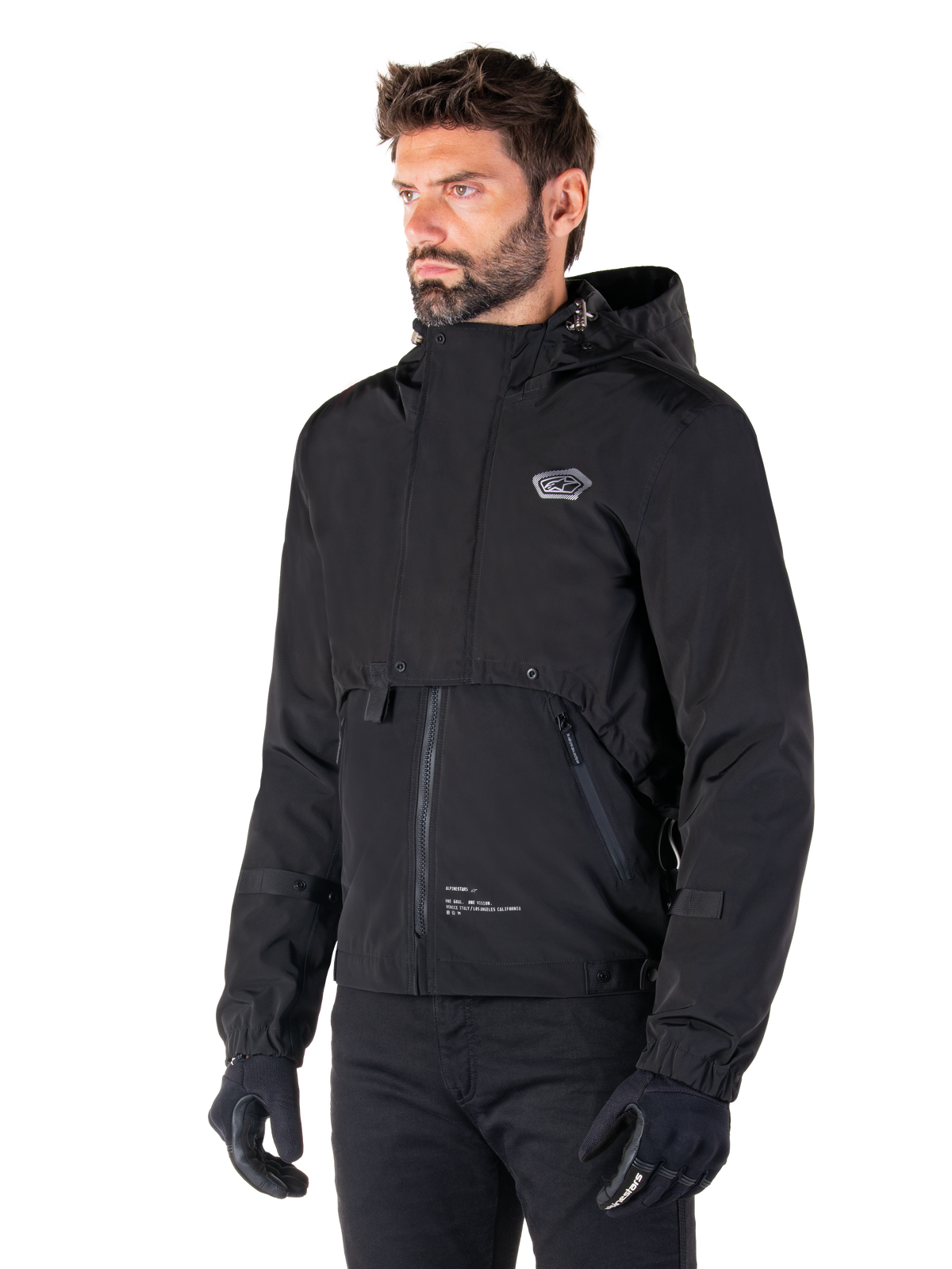 Alpinestars Parka Mootant Waterproof, giacca da moto modulare nera, indossata da un uomo con guanti e pantaloni neri, presenta cappuccio, cerniera frontale e design utilitaristico a strati, estetica urbana elegante con logo sul petto.