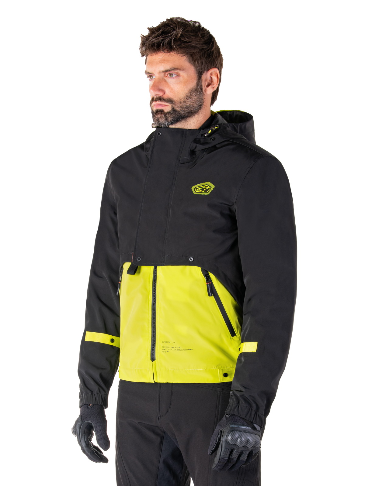 Alpinestars Mootant Waterproof Parka in Lime Punch Black, giacca da moto modulare indossata da un uomo con guanti e pantaloni da guida neri, caratterizzata da una sezione superiore e maniche nere con un pannello inferiore verde lime vivace, design con cappuccio dall'estetica utilitaristica di ispirazione militare.