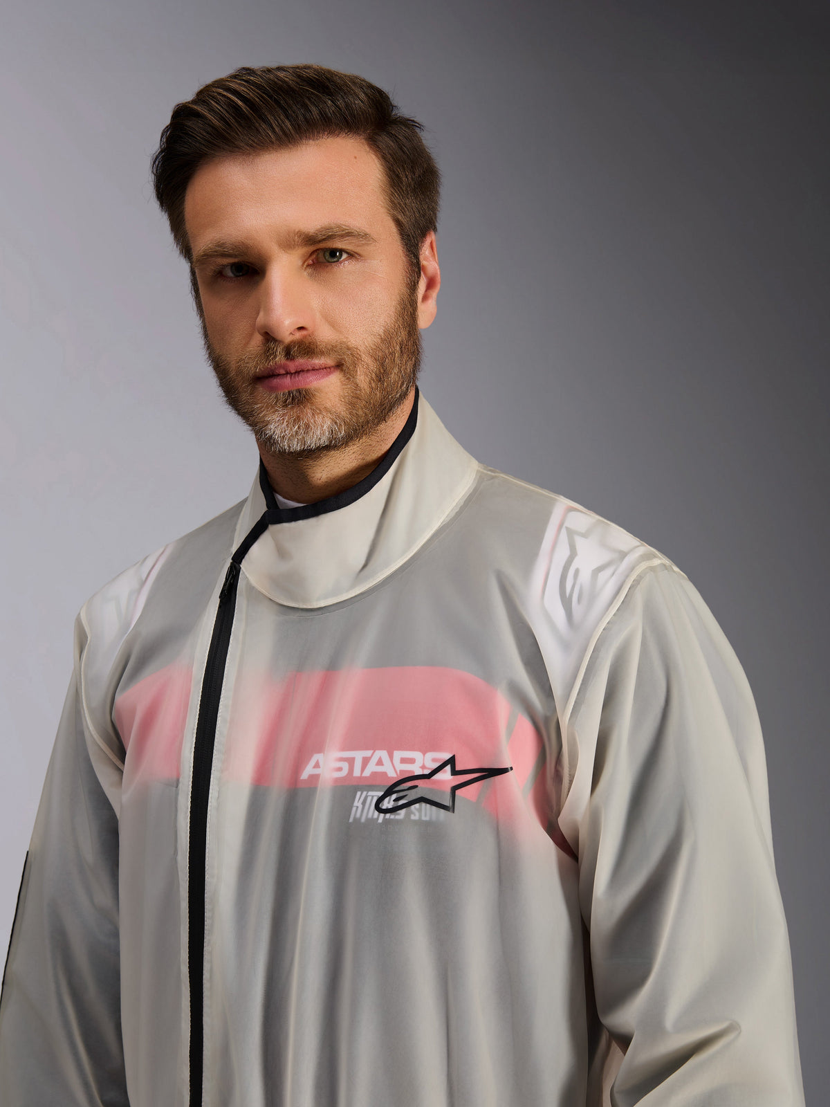 Alpinestars Kart Rain Suit, sovratuta antipioggia da kart, trasparente, indossata da un uomo, materiale impermeabile trasparente che mostra i dettagli della tuta da corsa rossa e bianca sottostante, collo alto con cerniera nera e logo a stella Alpinestars sul petto