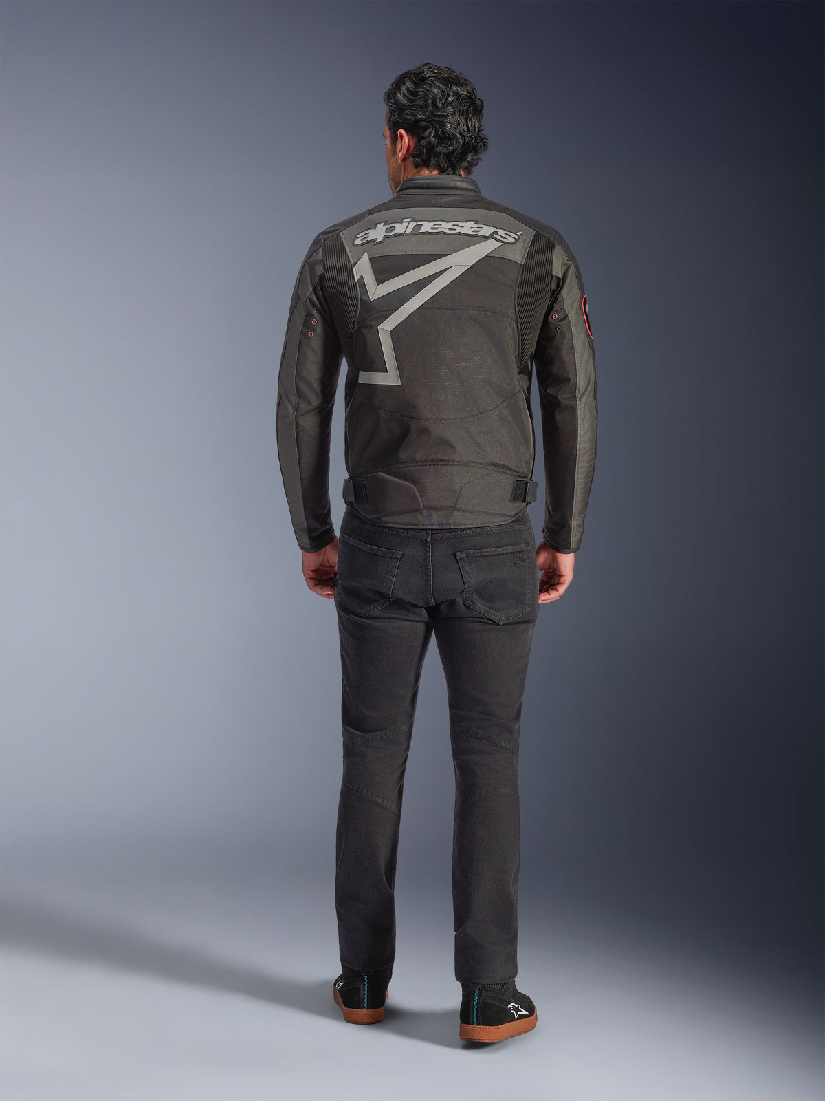 Giacca Alpinestars Axton, giacca da moto, Nero Antracite Rosso Tangerine, vista posteriore con grande logo Alpinestars grigio e grafica a stella, pannelli a fisarmonica sulle spalle, indossata con pantaloni neri e scarpe casual nere.