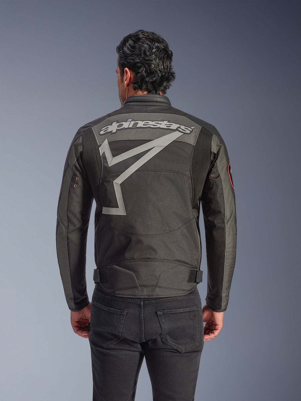 Alpinestars Giacca Axton, giacca da moto da uomo, nero antracite rosso tangerine, vista posteriore con grande marchio Alpinestars grigio e grafica a stella stilizzata, pannelli a fisarmonica sulle spalle e occhielli in metallo d'ispirazione vintage sulle maniche.