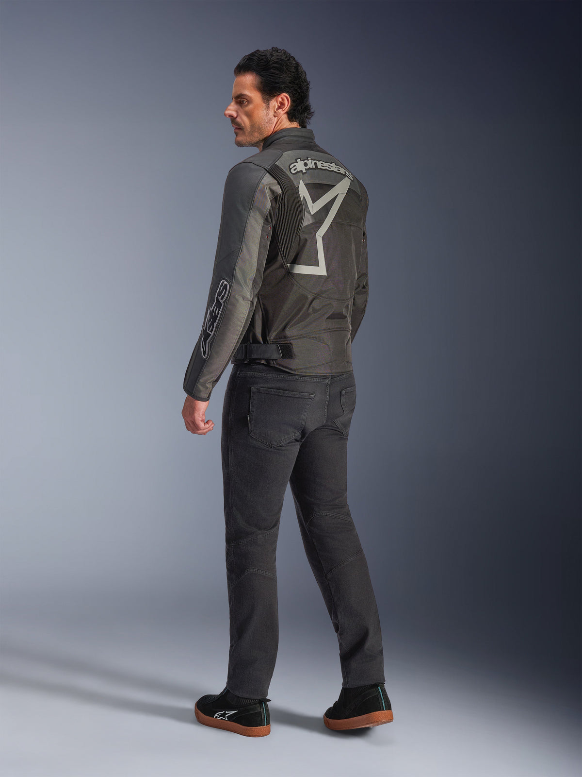Giacca Alpinestars Axton, giacca da moto da uomo, nero e antracite, vista posteriore con un grande logo grigio avvolgente e pannelli a fisarmonica sulle spalle, indossata con pantaloni da guida scuri e sneakers nere, stile urban contemporaneo con eredità racing