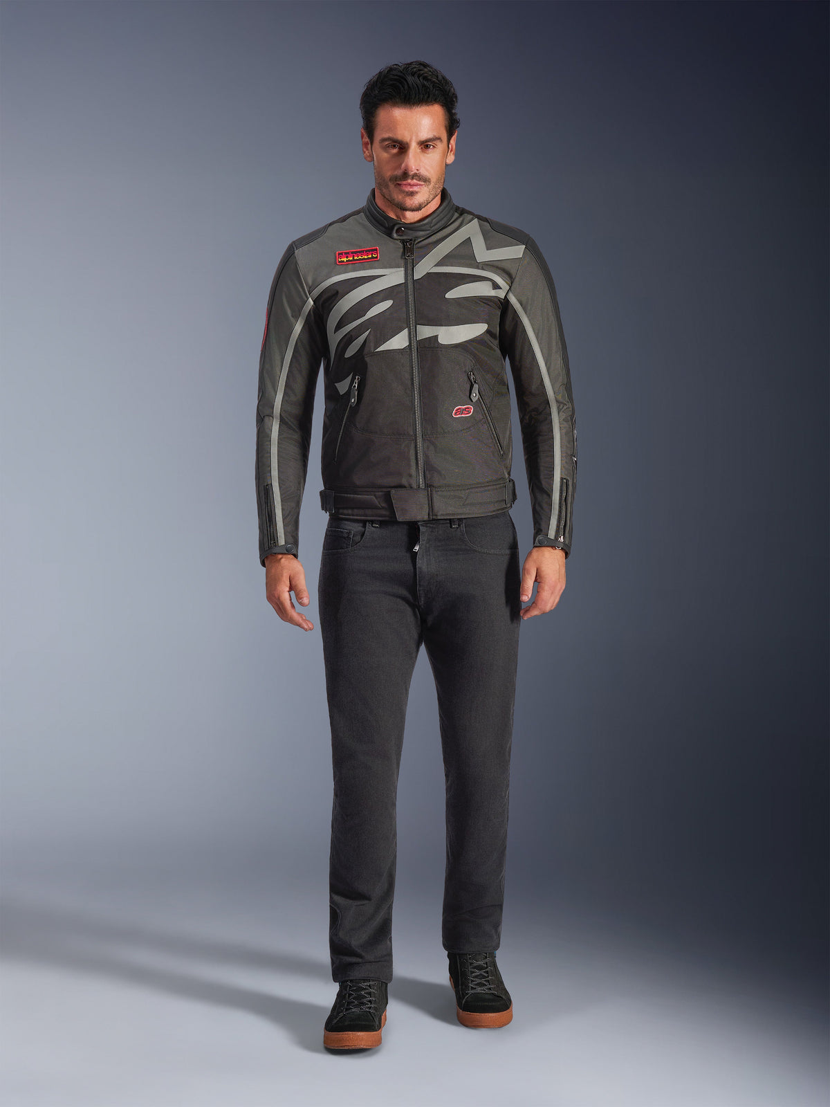 Giacca Alpinestars Axton, giacca da moto, nero antracite rosso tangerine, indossata da un modello con pantaloni scuri, con logo Alpinestars grigio avvolgente, patch del marchio rosse e guscio in tessuto resistente all'acqua per la guida urbana