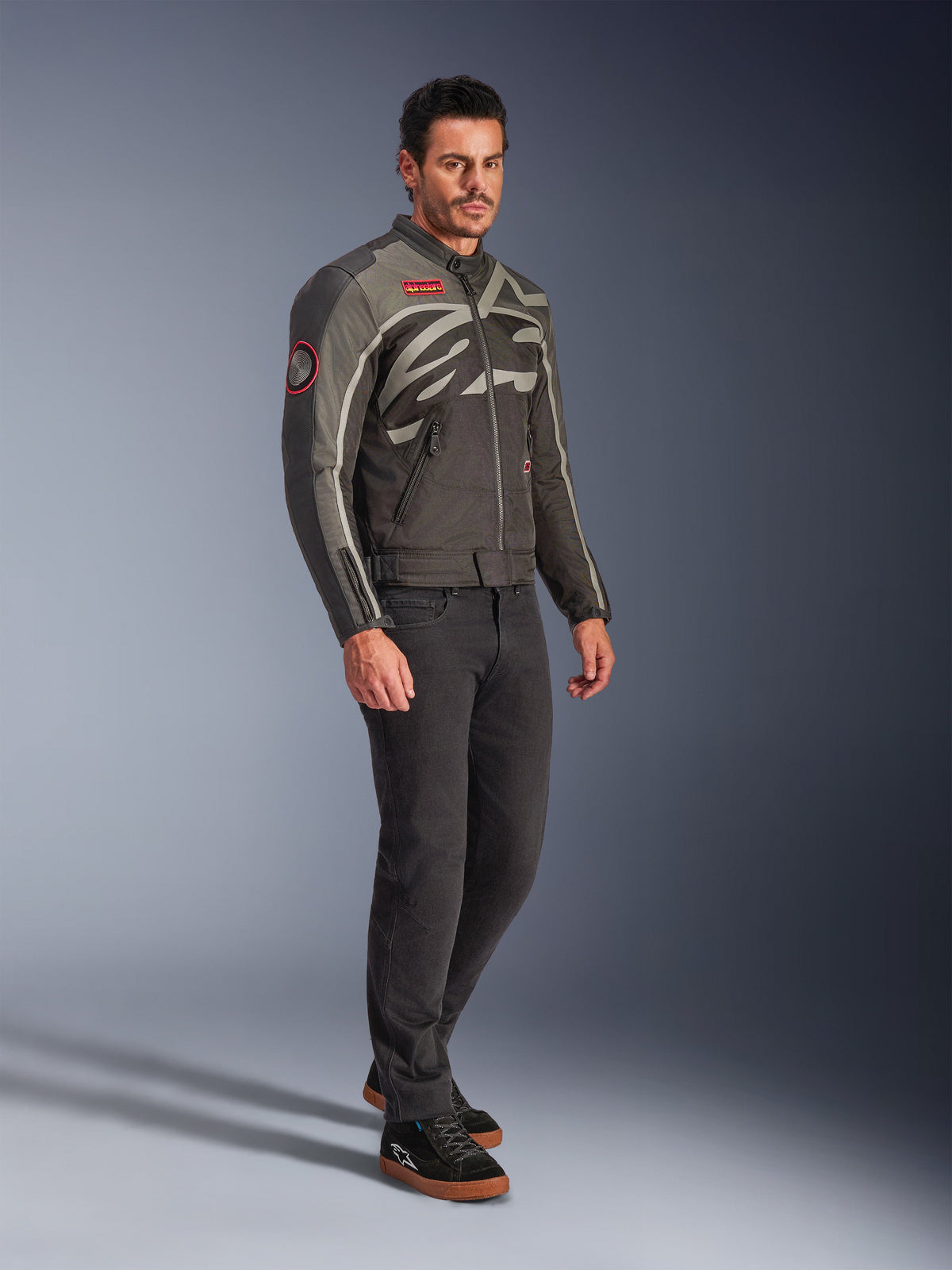 Modello che indossa la Giacca Alpinestars Axton in Nero Antracite Rosso Tangerine, giacca da moto d'ispirazione vintage con logo avvolgente e dettagli rossi, abbinata a pantaloni neri e sneaker da moto nere, abbigliamento da moto urban contemporaneo