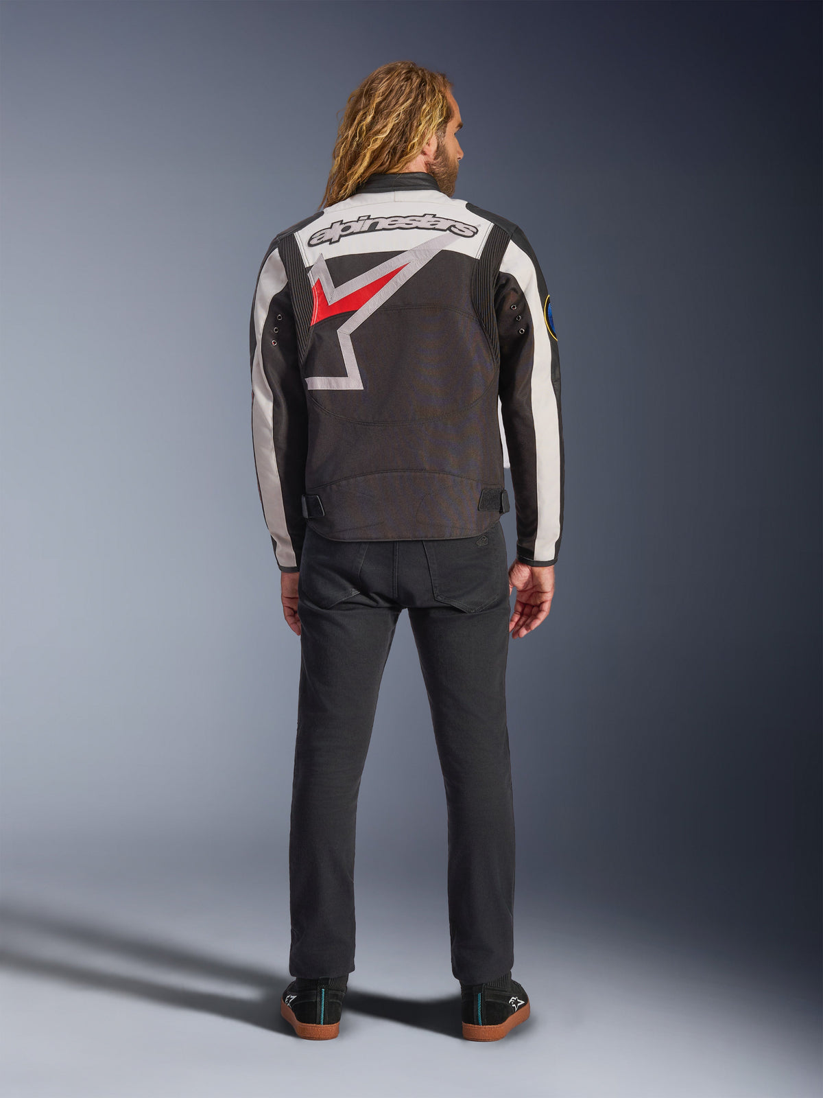 Giacca Alpinestars Axton, giacca da moto, nero, bianco sporco e rosso brillante, vista posteriore su un modello, con un grande logo a stella Alpinestars sul retro, pannelli delle maniche bianco sporco, indossata con pantaloni neri, design ispirato all'abbigliamento racing vintage