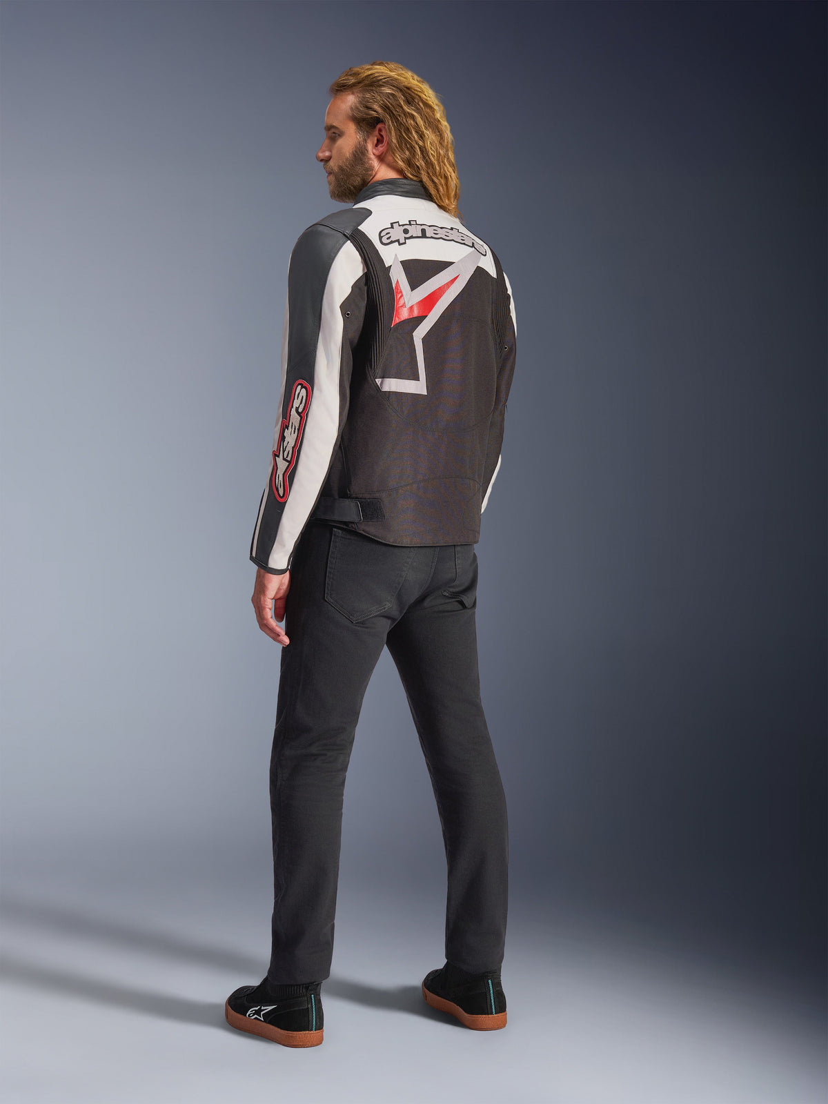 Giacca Alpinestars Axton, giacca da moto, nero, bianco sporco e rosso acceso, vista posteriore indossata da un modello, con grande logo a stella Alpinestars e scritta sulla schiena, design d'ispirazione racing vintage con pannelli a fisarmonica, abbinata a pantaloni neri e sneakers
