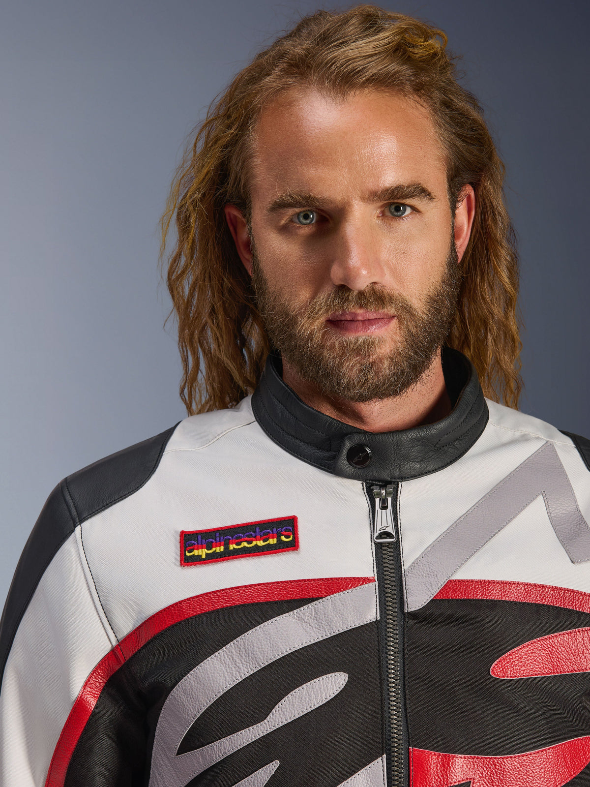 Uomo che indossa la giacca Alpinestars Axton, giacca da moto d'ispirazione vintage in nero, bianco sporco e rosso vivace, vista ravvicinata della toppa sul petto e del colletto minimalista, abbigliamento da moto contemporaneo