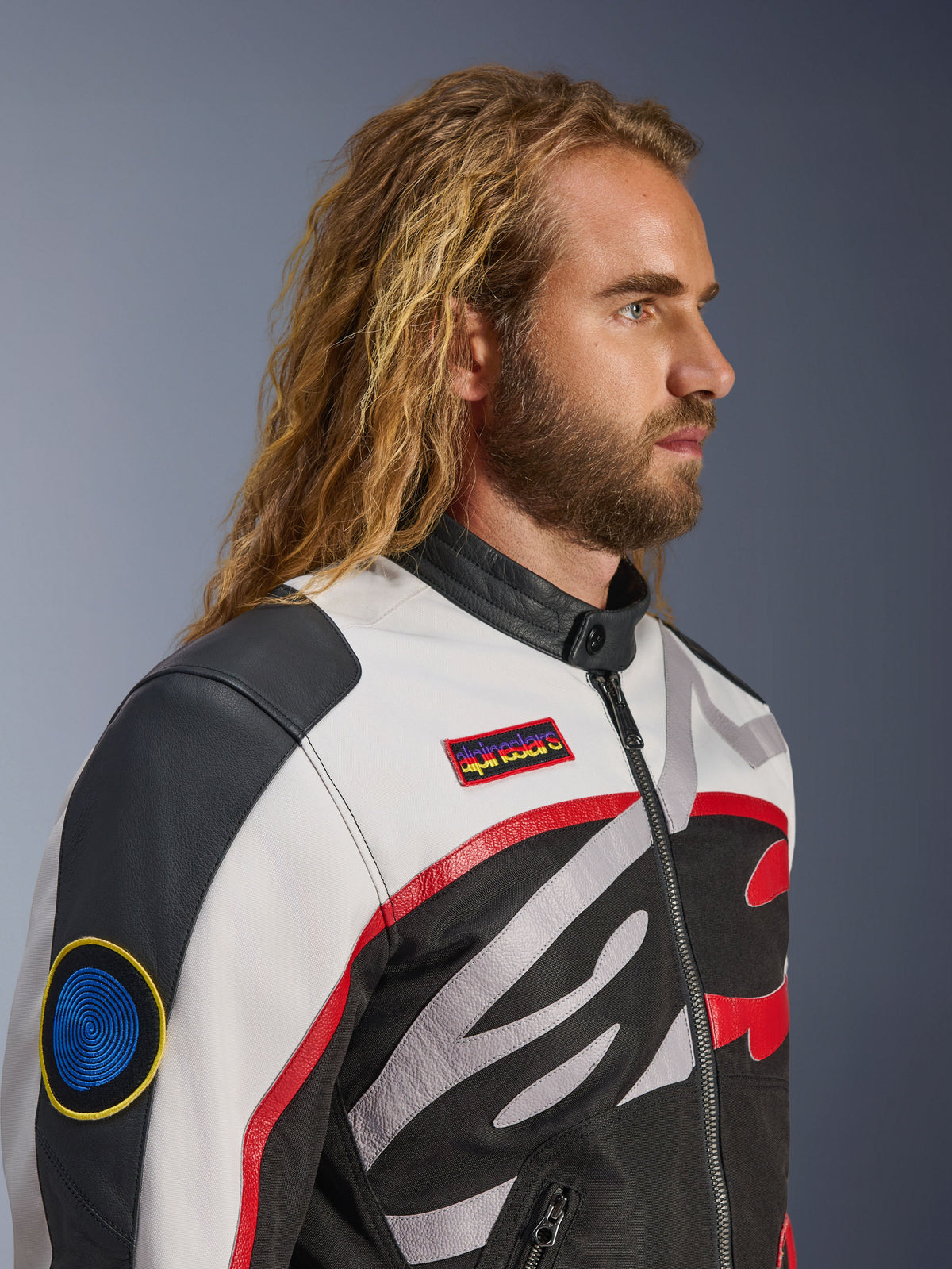Uomo con capelli lunghi e barba che indossa la Giacca Alpinestars Axton, giacca da moto, nero, bianco sporco e rosso vivace, design ispirato al racing vintage con patch logate e logo avvolgente, vista di profilo ravvicinata, stile contemporaneo urbano