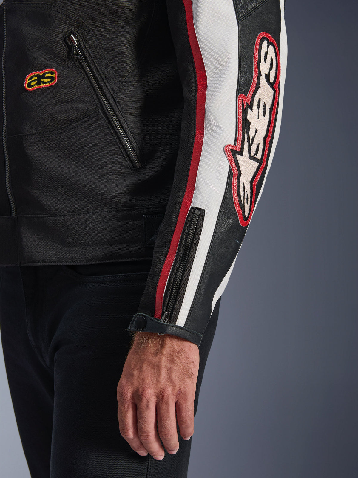 Giacca Alpinestars Axton, primo piano di manica e busto, nero con dettagli bianco panna e rosso vivace, grande logo Alpinestars ricamato sul braccio, strisce verticali stile racing, polsino con zip e bottone automatico, robusta struttura in tessuto