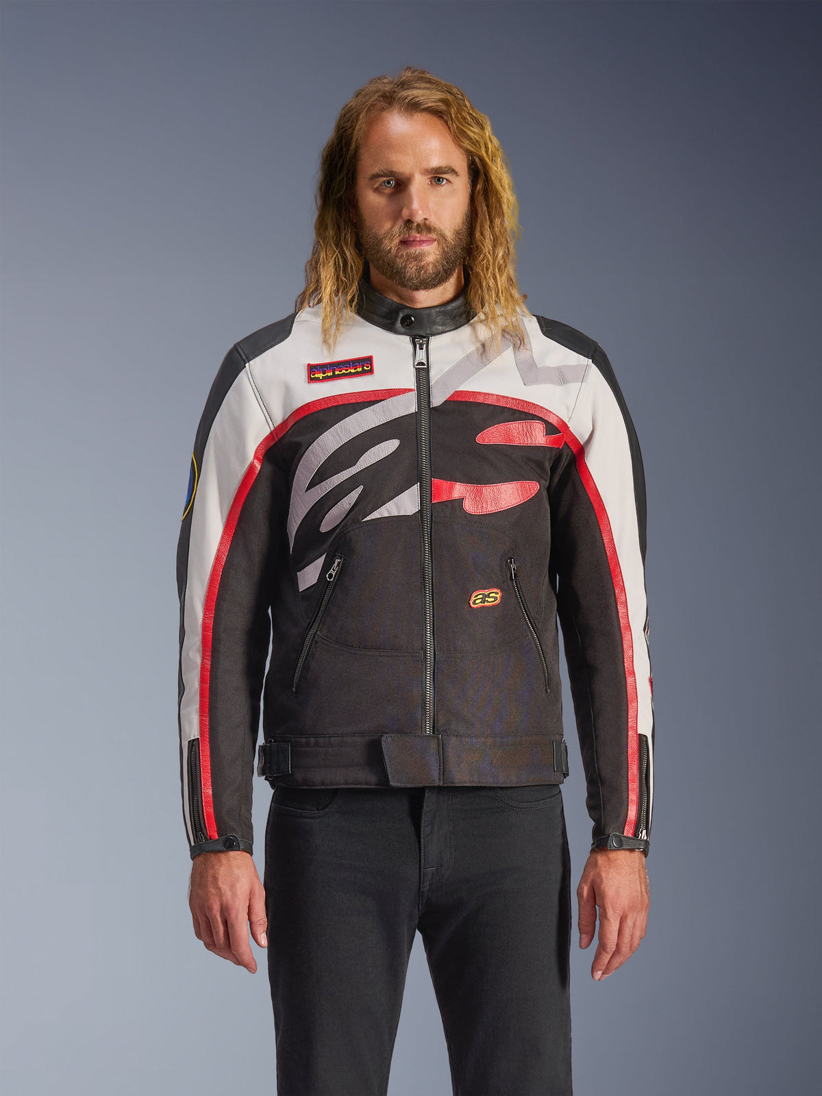Giacca Alpinestars Axton, giacca da moto, nero, bianco panna e rosso vivace, con audaci motivi astratti sul petto e logo avvolgente, maniche con strisce rosse, design di ispirazione vintage con colletto minimalista, indossata da un modello con jeans neri