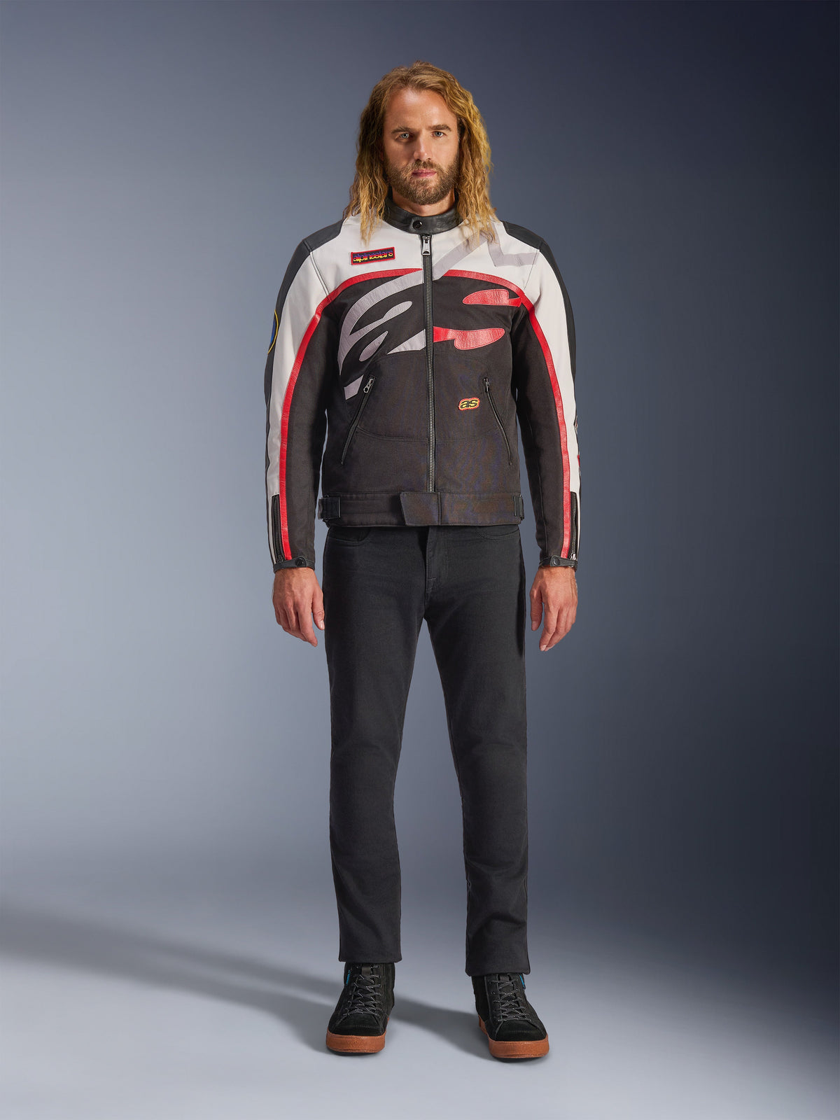 Giacca Alpinestars Axton, giacca da moto, nero, bianco sporco e rosso acceso, indossata da un modello con pantaloni neri e sneakers, con un audace logo avvolgente e un design d'ispirazione racing vintage
