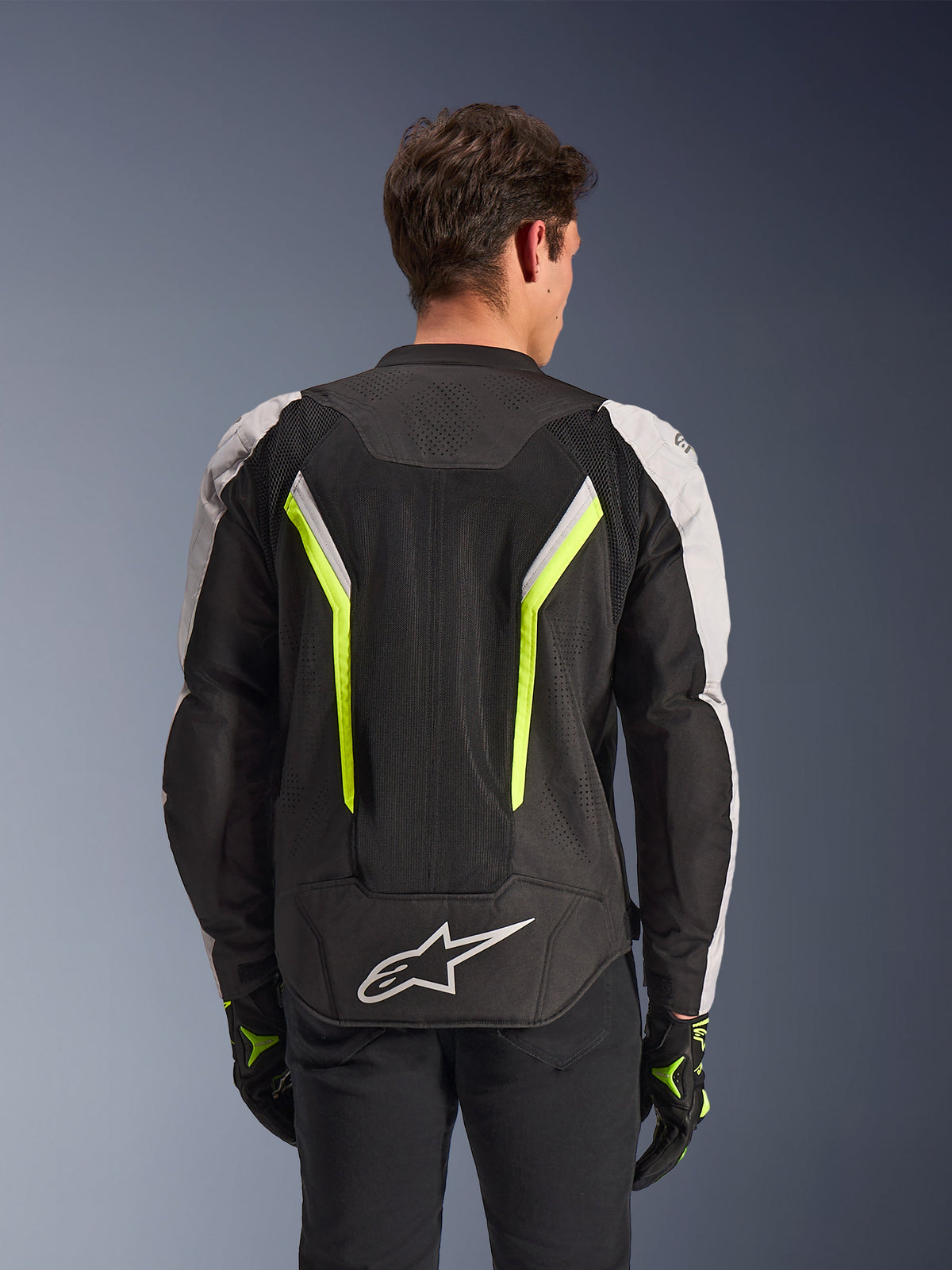 Alpinestars AST V3 Air Jacket, Black Silver Yellow Fluo, vista posteriore su modello, giacca da moto in tessuto con pannelli di ventilazione in doppia rete tecnica, strisce verticali giallo fluo, maniche argento e logo Alpinestars bianco sulla parte bassa della schiena
